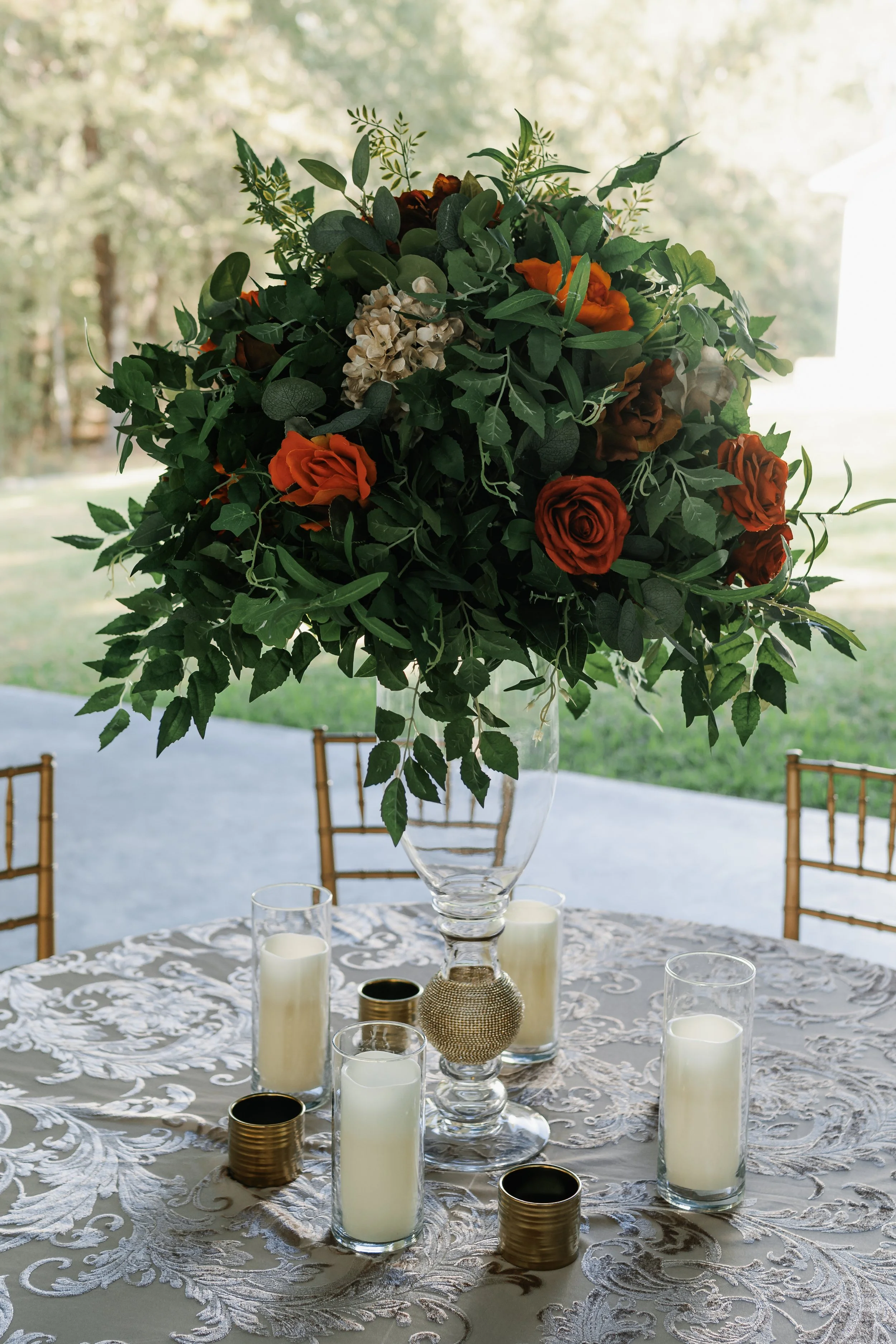 A floral centerpiece with red roses, white hydrangeas, and greenery on a table with acrylic candle holders and candles, set outdoors with a grassy background.