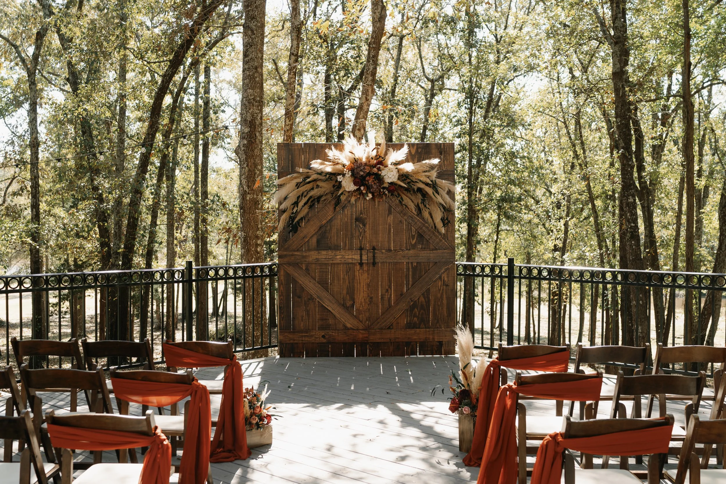 Outdoor wedding setup with wooden chairs decorated with orange sashes, facing a wooden backdrop with floral arrangements, set in a wooded area.