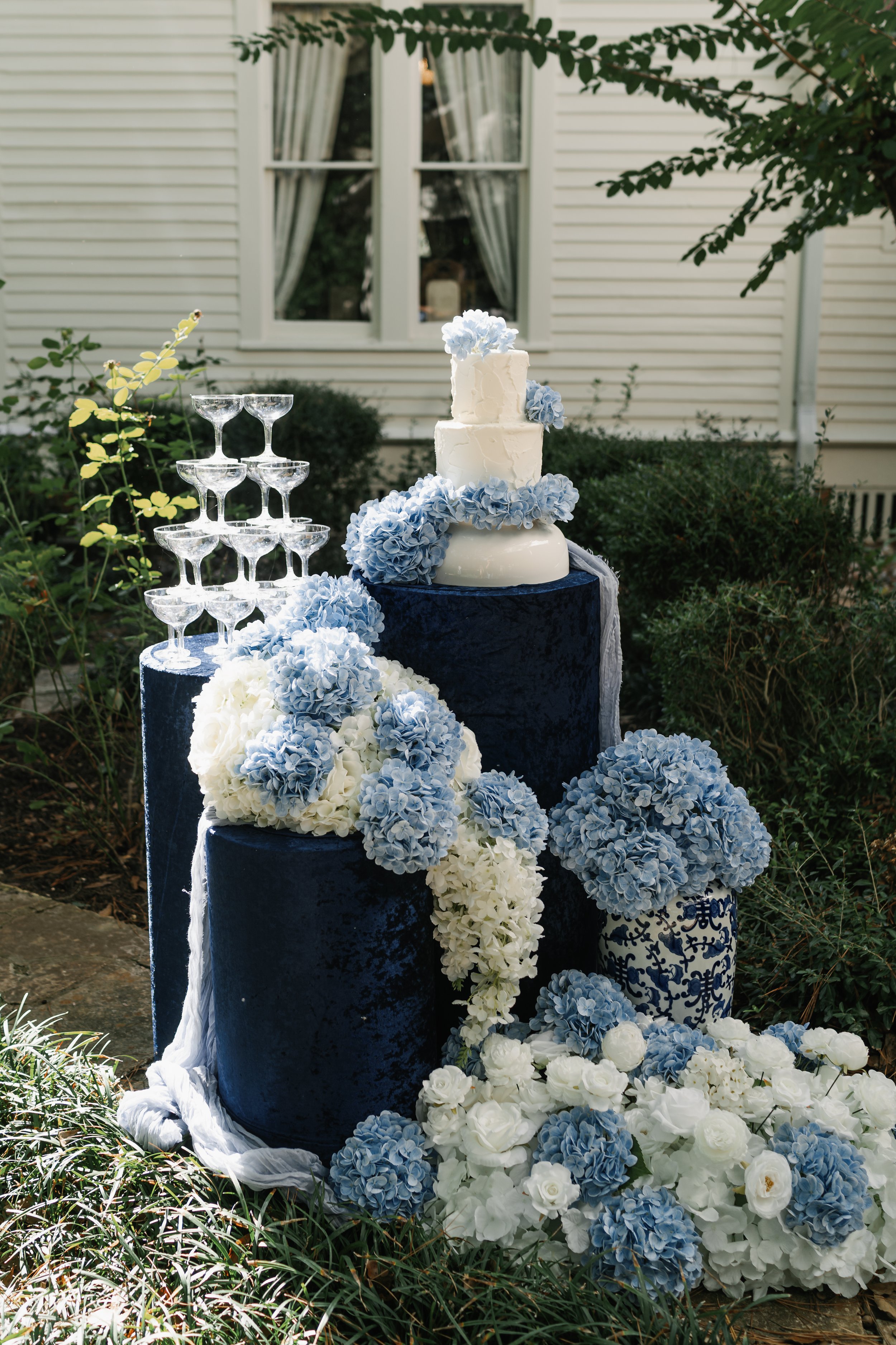 A wedding cake and floral arrangement outdoors, with a tiered cake topped with blue flowers, surrounded by white and blue hydrangeas and roses, and a display of stacked coupe glasses on a dark blue velvet table.