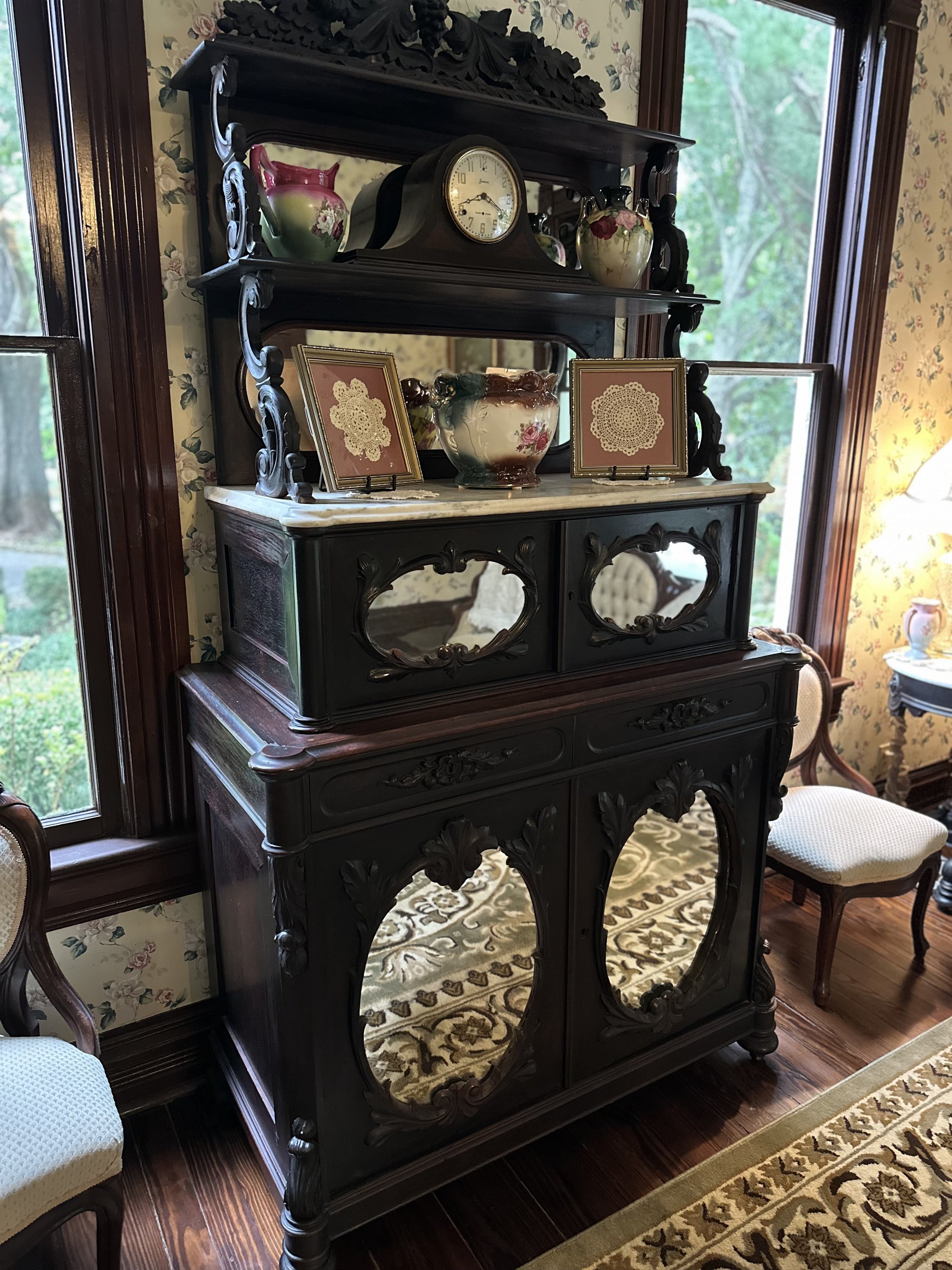 An antique black wooden cabinet with ornate carvings and mirrors, topped with decorative plates, doilies, and vases, situated in a room with floral wallpaper and hardwood floors, near a window with a view of trees outside.