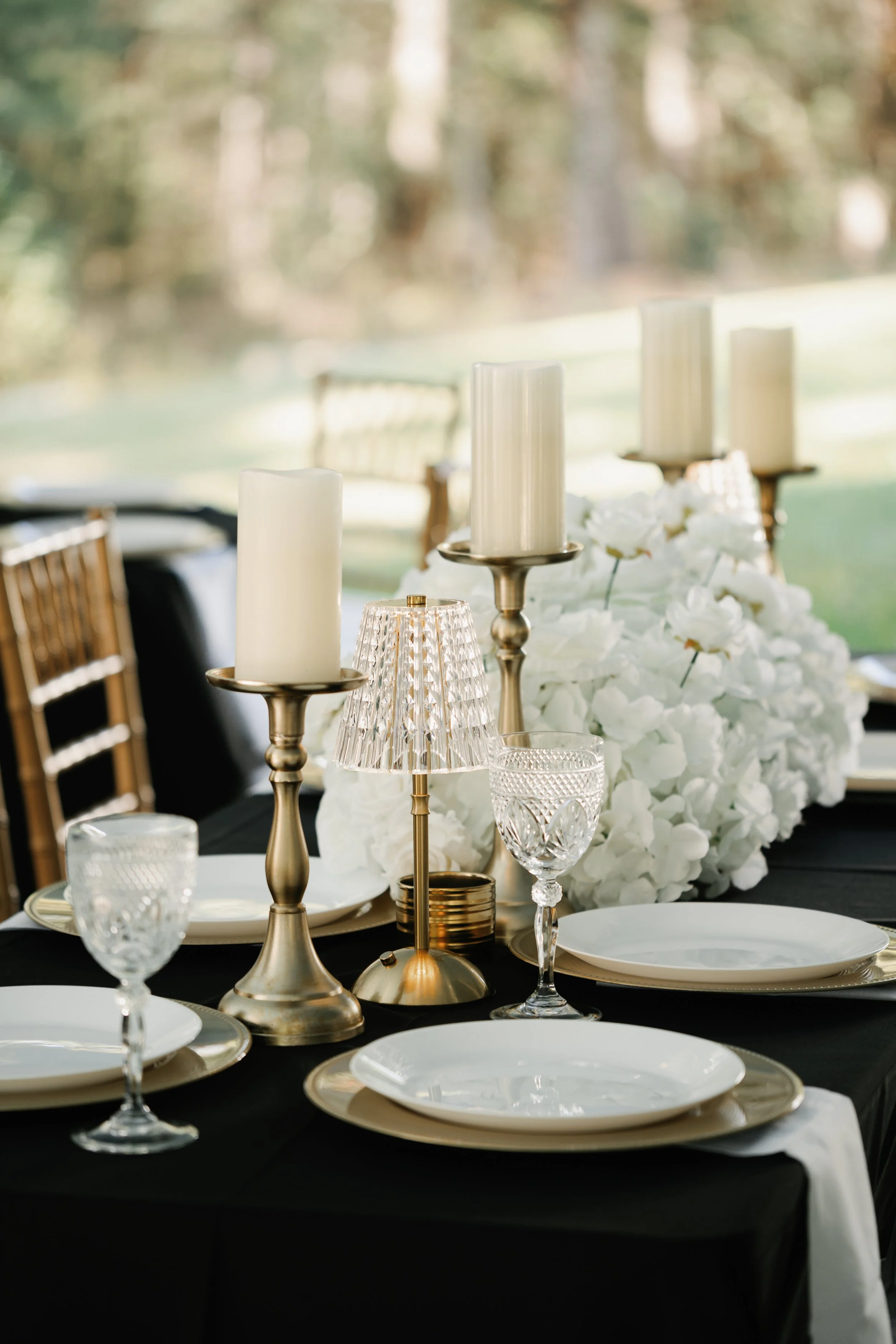 Elegant table setting with gold and crystal candlesticks, glassware, white plates, and a large floral centerpiece of white flowers at an outdoor event.