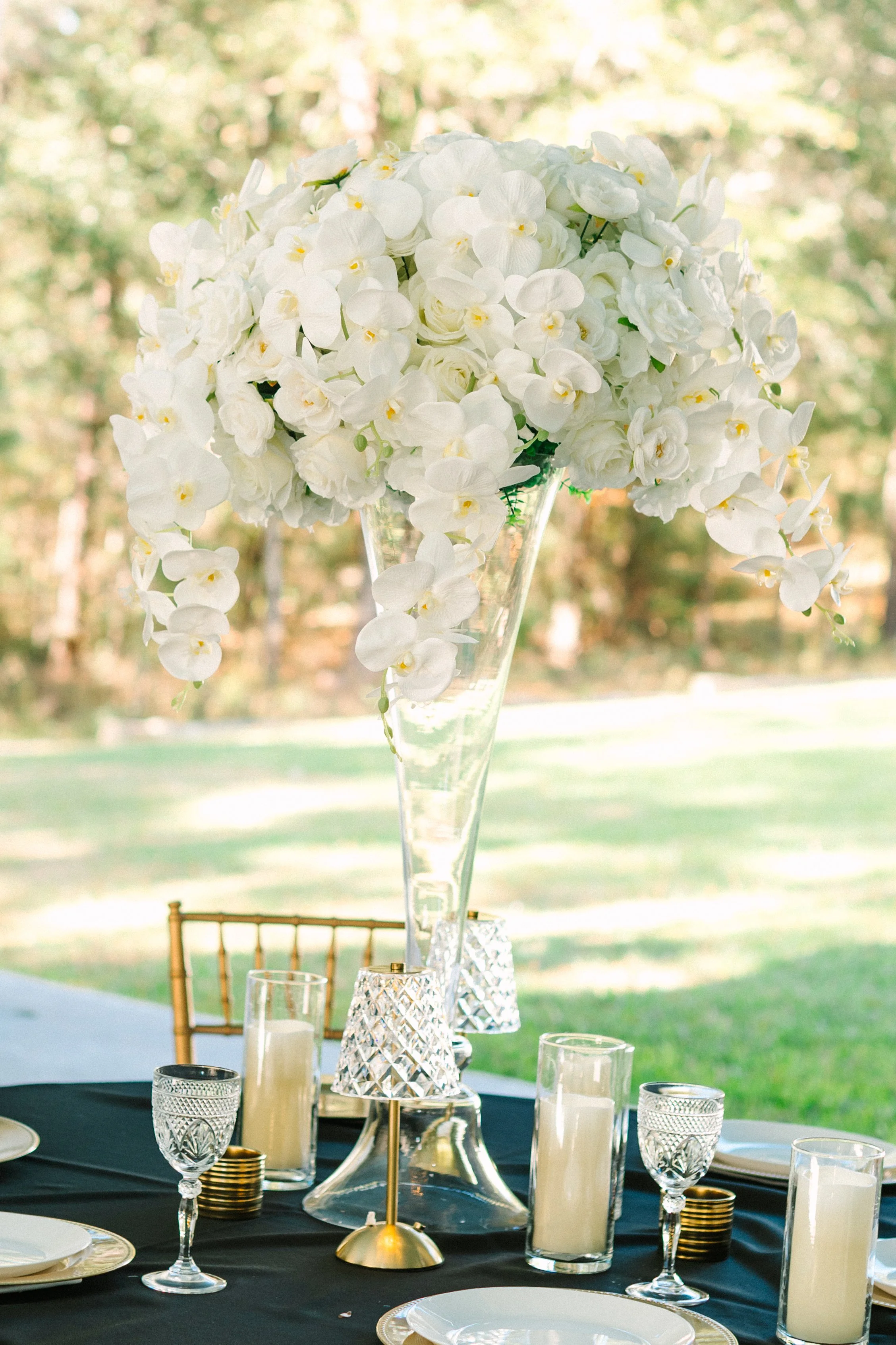 Elegant outdoor dining table with a tall floral centerpiece of white orchids and roses in a clear glass vase, black tablecloth, gold and crystal glassware, and candles, in a sunny garden setting.