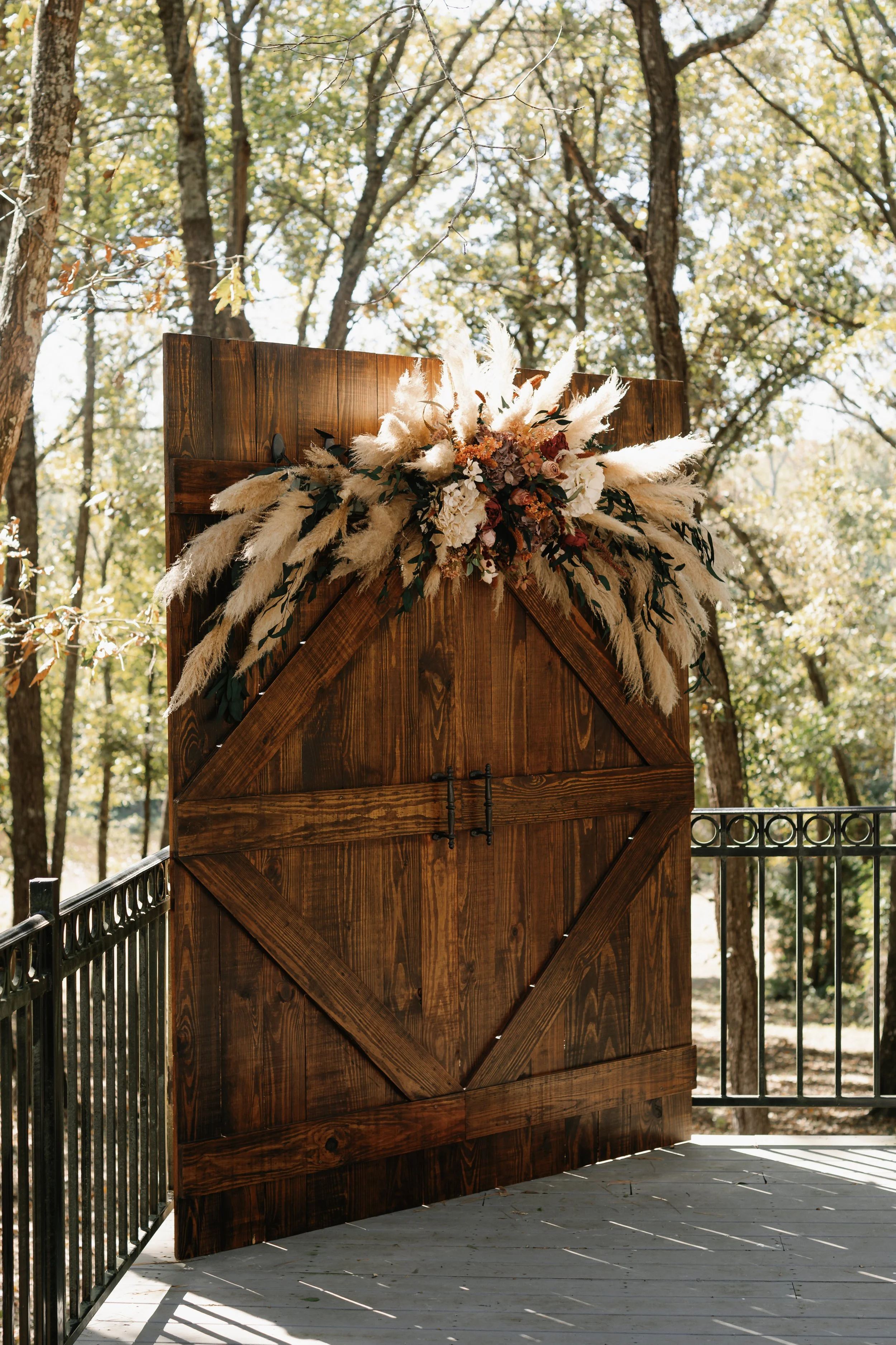 A large wooden barn door decorated with a floral arrangement of white pampas grass, dried flowers, and greenery, set outdoors surrounded by trees.