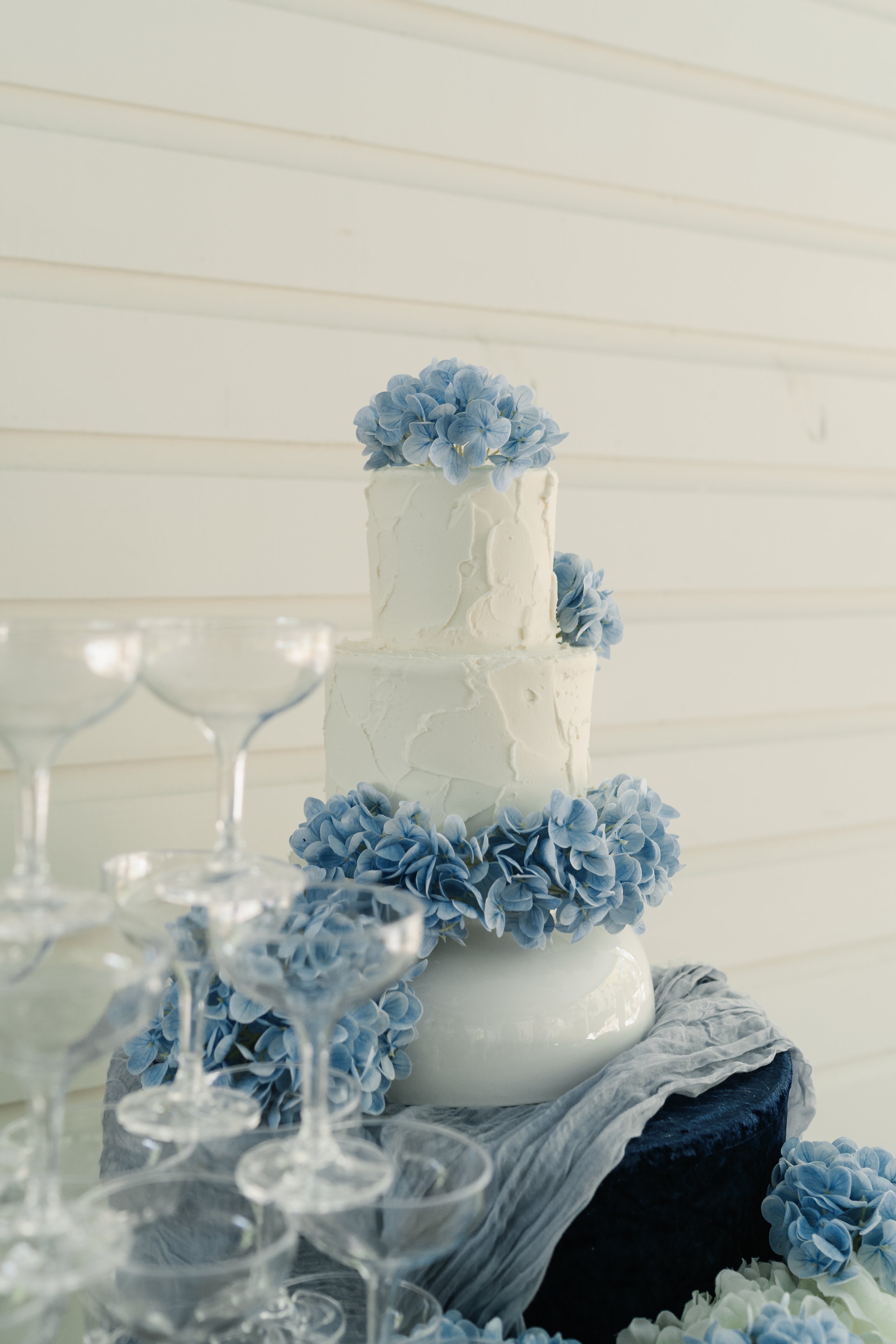 Two-tier white wedding cake decorated with light blue hydrangea flowers, surrounded by empty coupe glasses on a table with dark fabric and gray cloth.