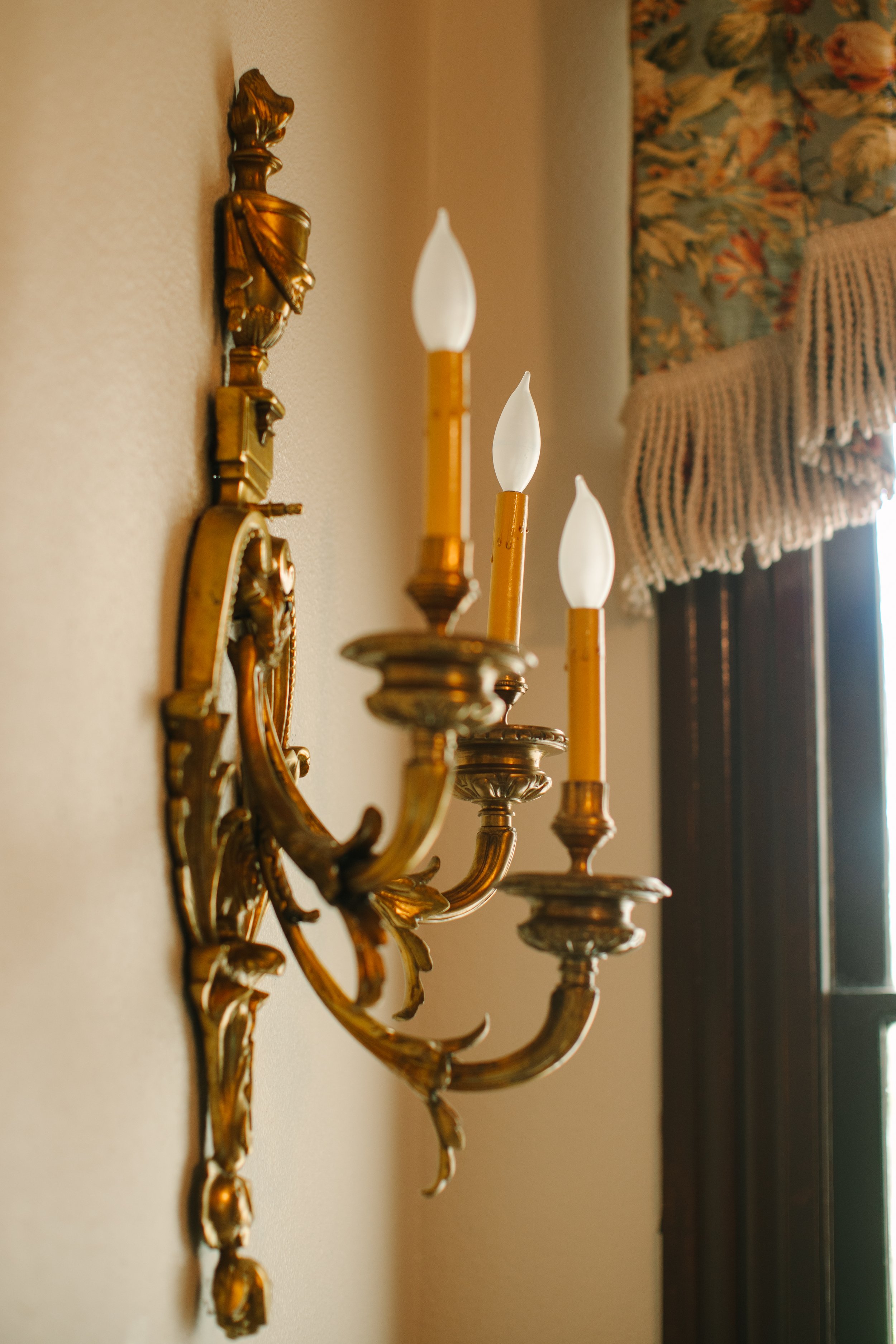 A vintage gold wall sconce with three candle-shaped bulbs, mounted on a beige wall near a window with floral curtains.