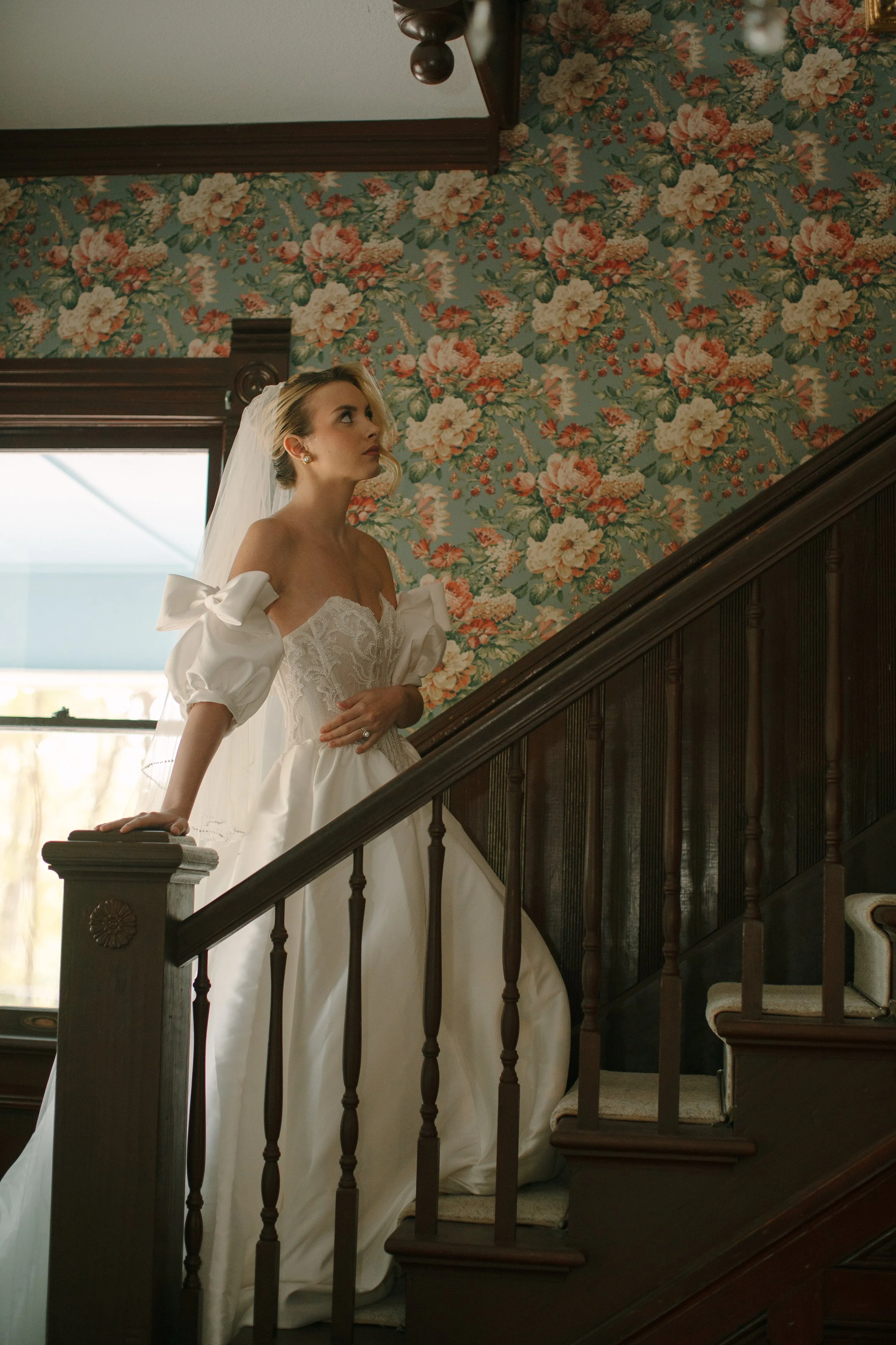 A bride in a white wedding gown with puffed sleeves and a veil stands on a wooden staircase with floral wallpaper in the background.