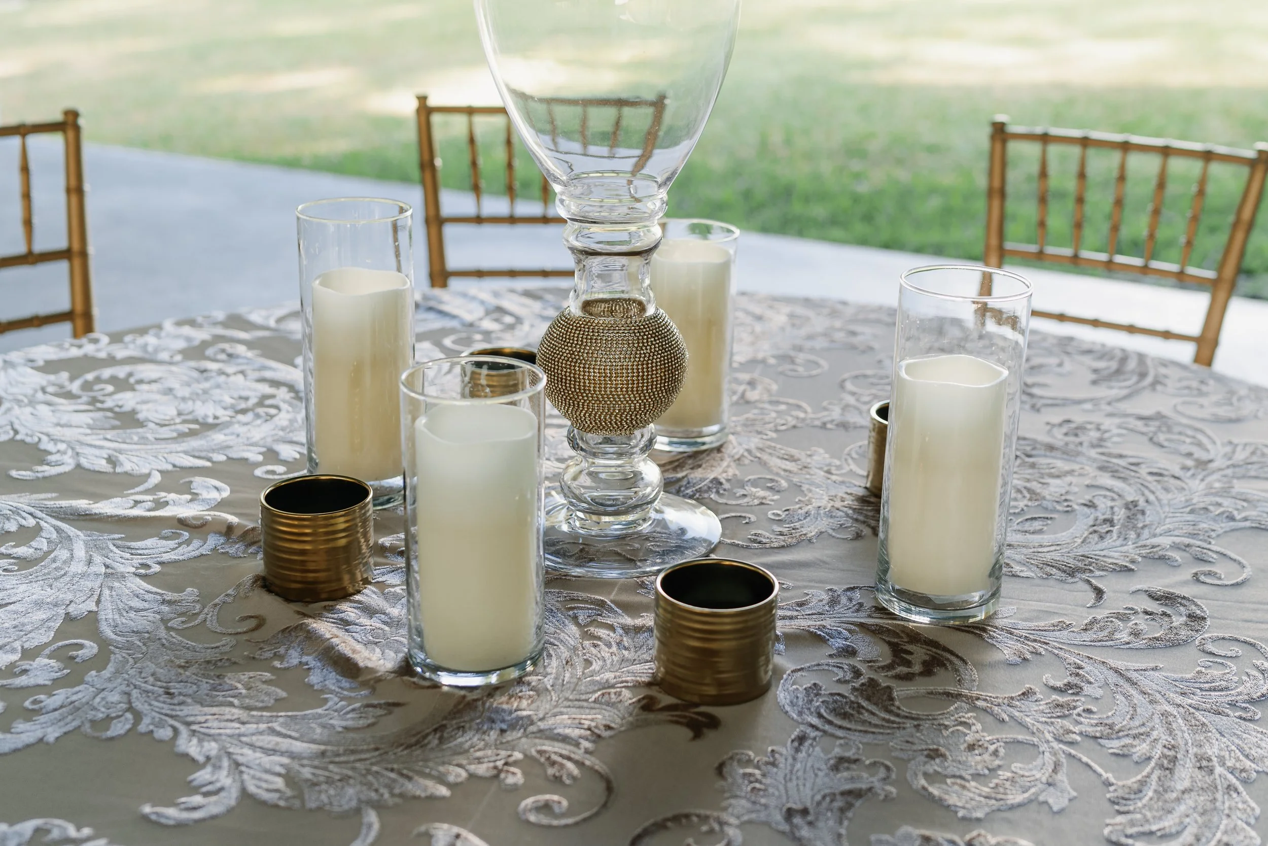 Elegant table setting with a white lace tablecloth, tall glass candle holders with white candles, a decorative glass and gold centerpiece, and gold holders, set outdoors with chairs and green lawn in background.
