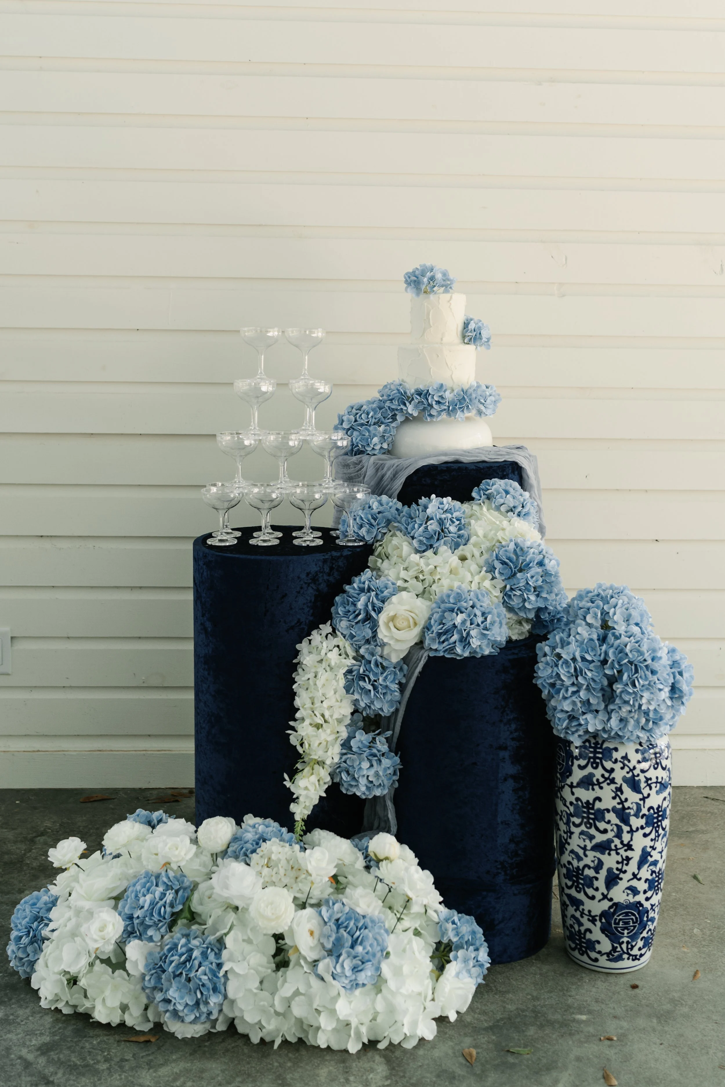 Decorative arrangement with a tiered white cake decorated with blue flowers, glassware, and blue and white floral arrangements on and around dark velvet pedestals against a white paneled wall.