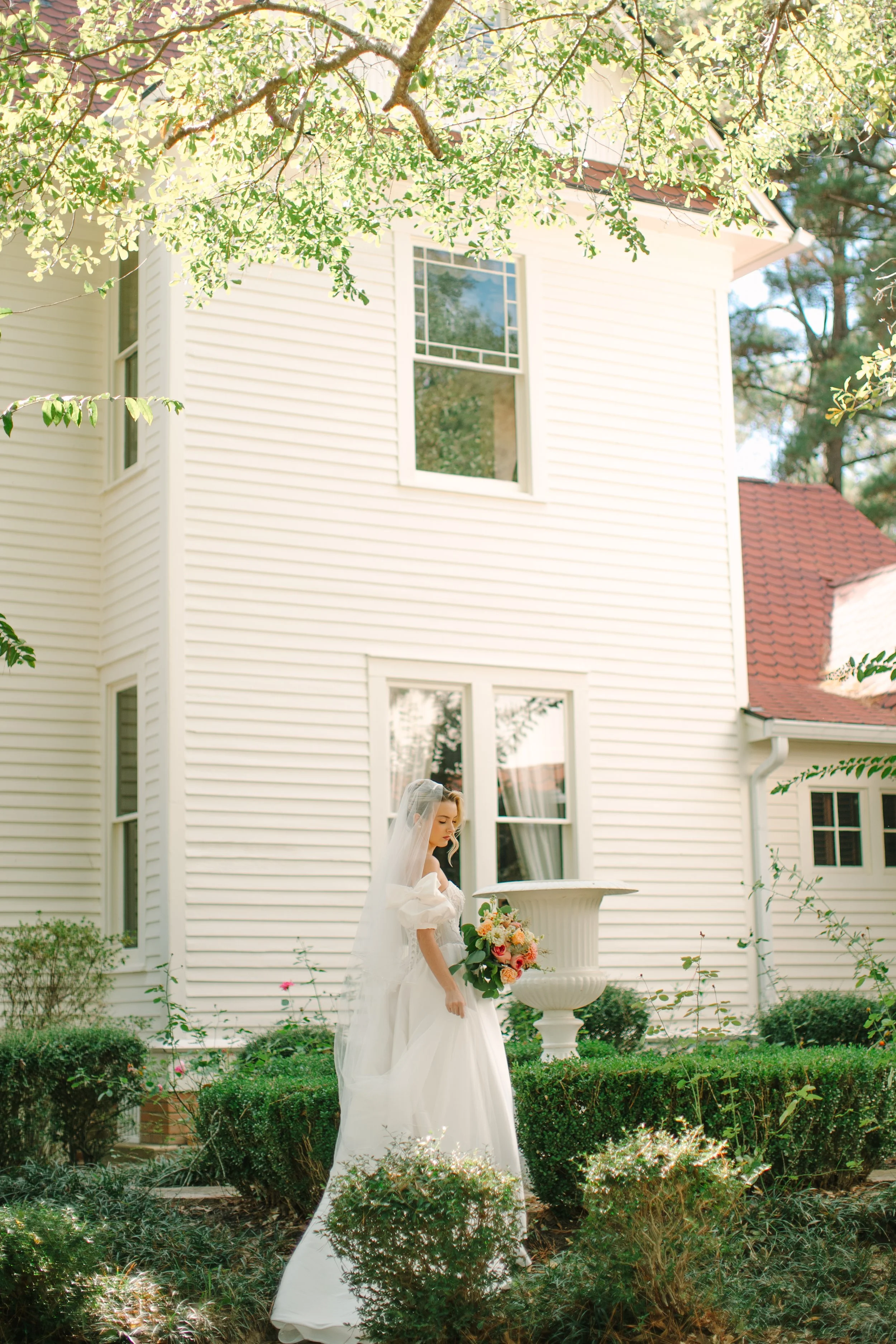 A bride in a white wedding gown and veil stands outdoors holding a bouquet of pink, peach, and white flowers, with a white house and lush green garden in the background.