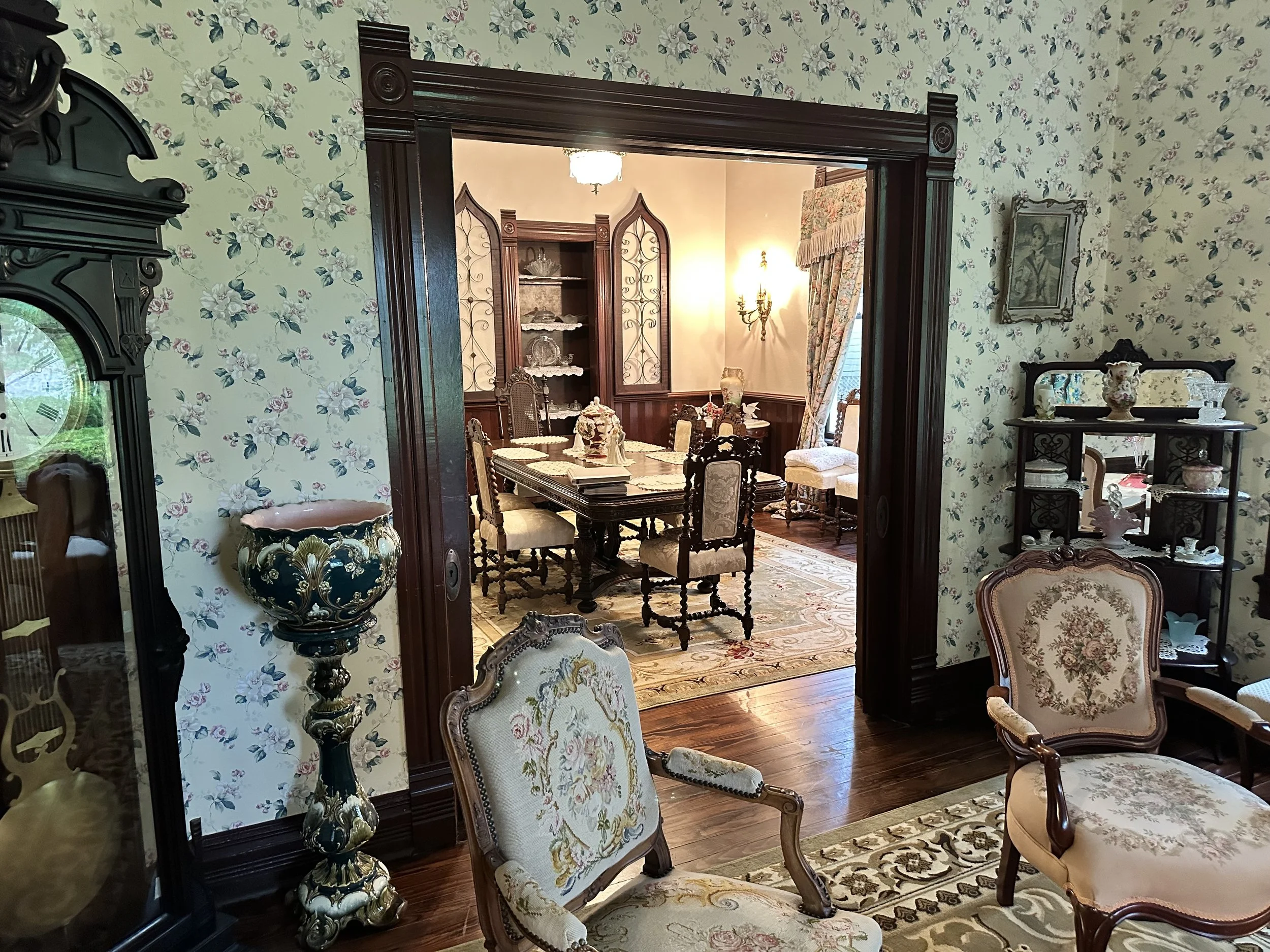 A view of a formal dining room and living room with vintage furniture, floral wallpaper, and decorative china shelves.