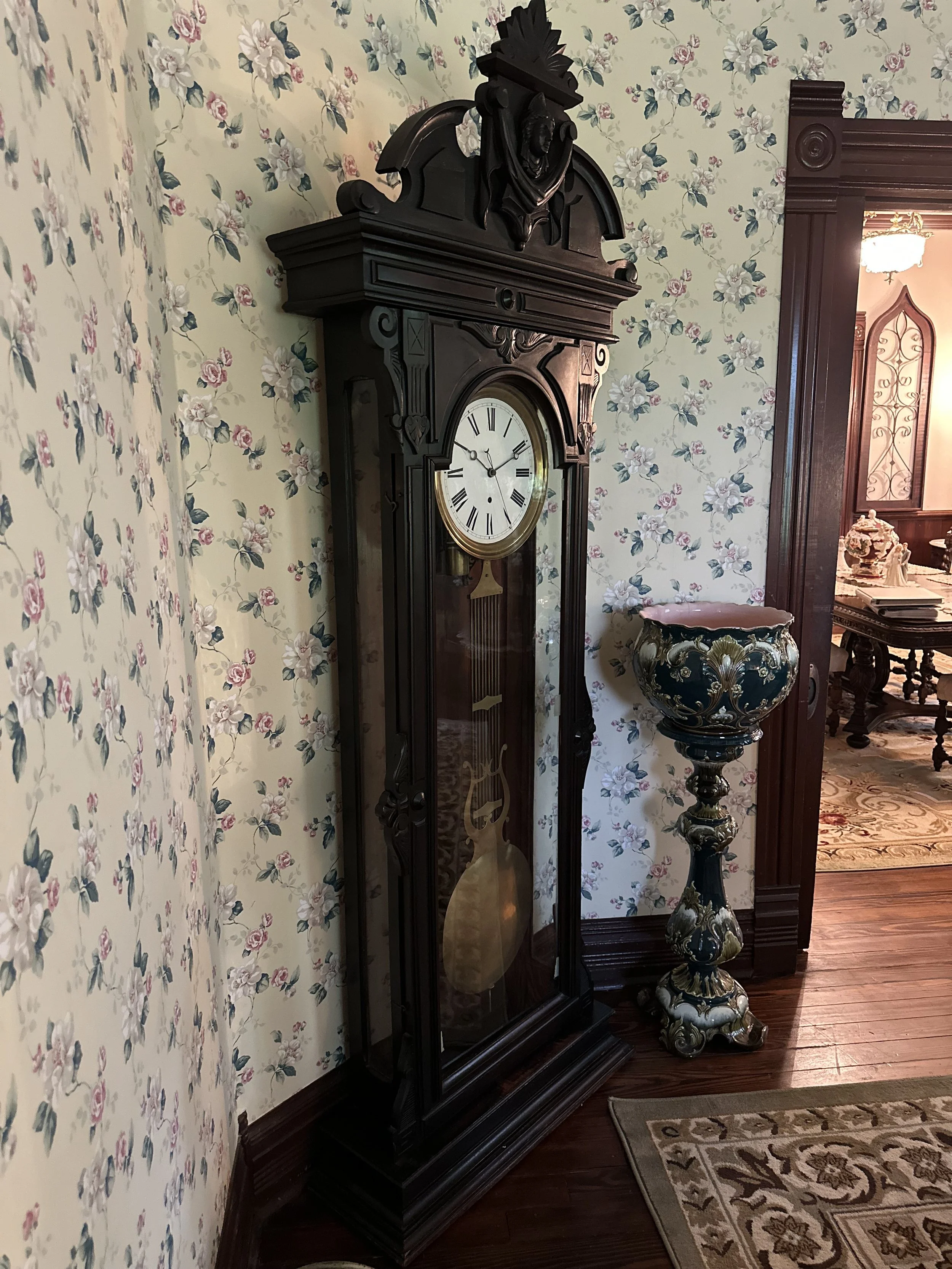An antique wooden grandfather clock with a white face and Roman numerals, standing next to a decorative pedestal with floral patterns, in a room with floral wallpaper and hardwood floors.