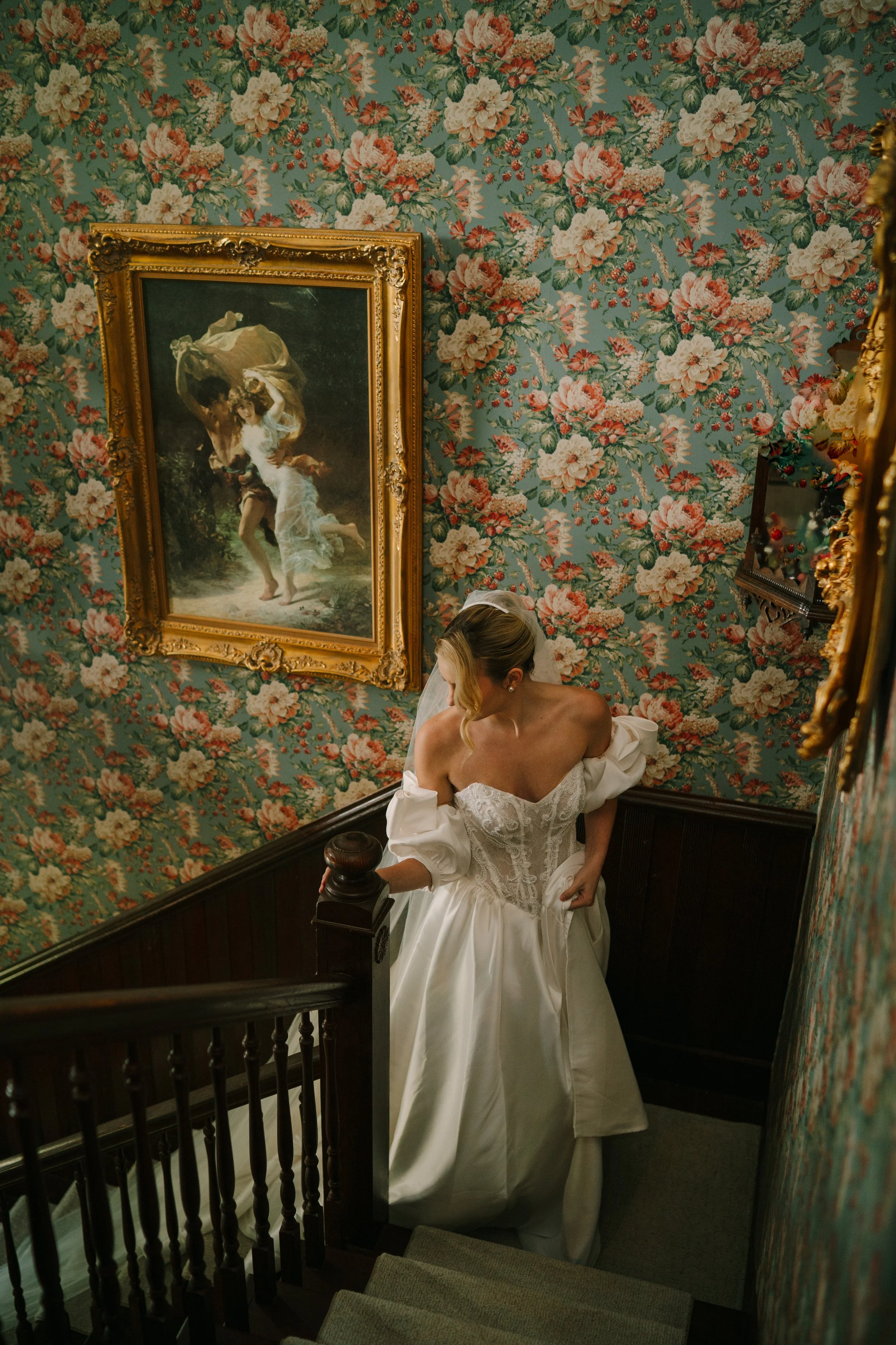 A woman in a wedding dress descending a staircase in a vintage decorated house with floral wallpaper and framed classical artwork.