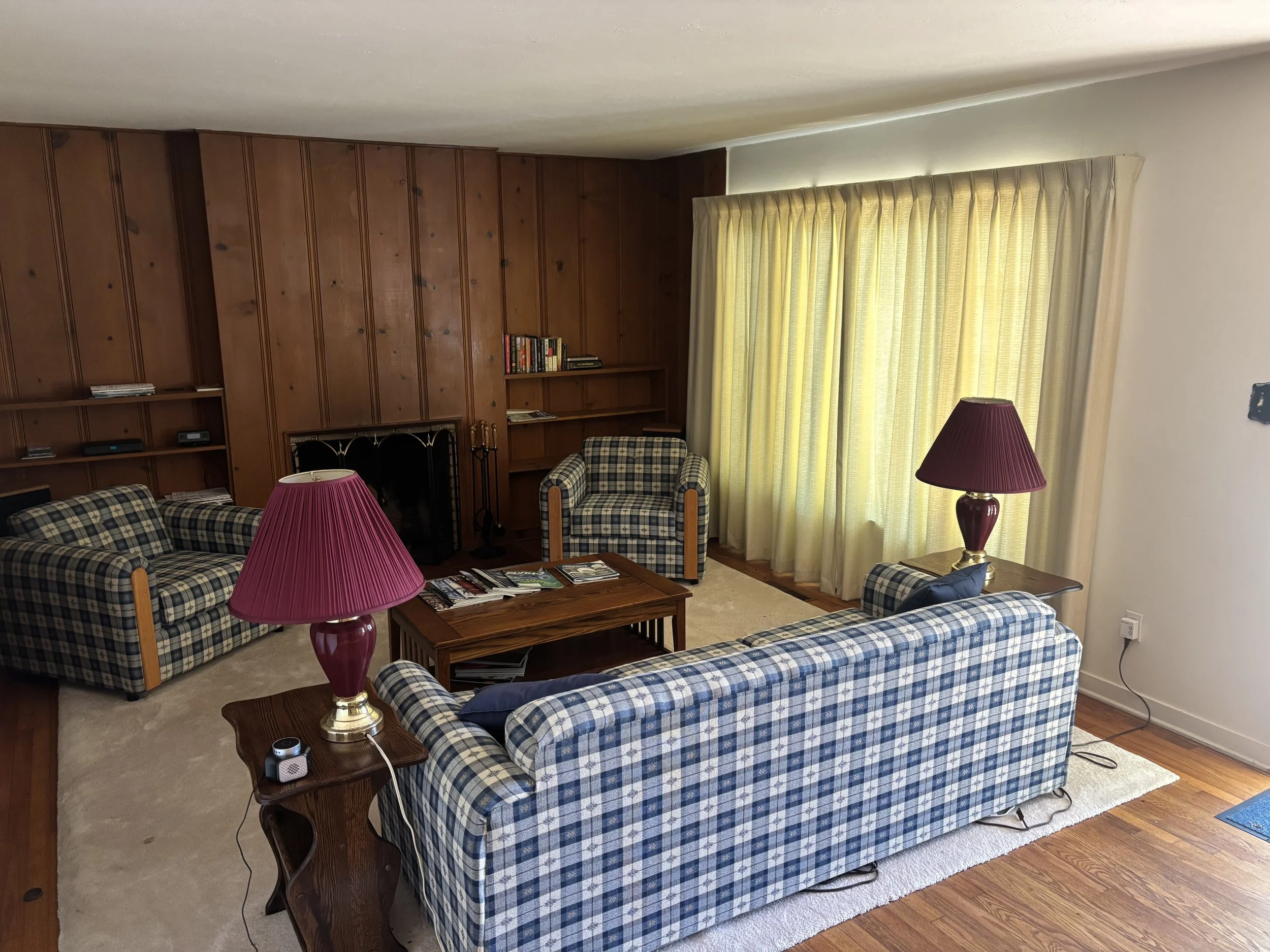 Living room with plaid sofas, wooden coffee table, lamp, bookshelf, and large window with yellow curtains.