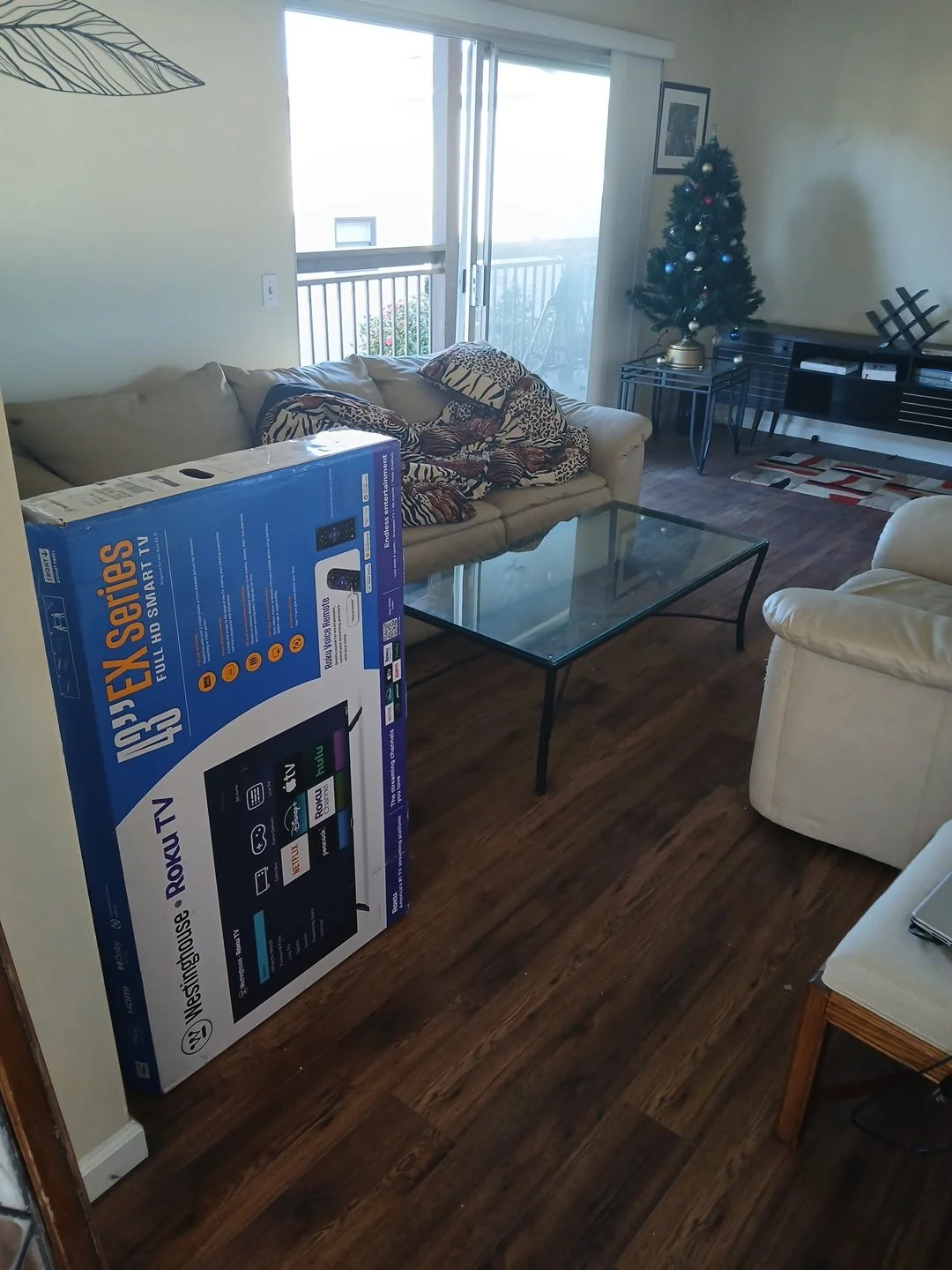 Living room with a couch wrapped with a blanket, a glass coffee table, a Christmas tree, a sliding glass door with a balcony, and a box for a Roku TV.