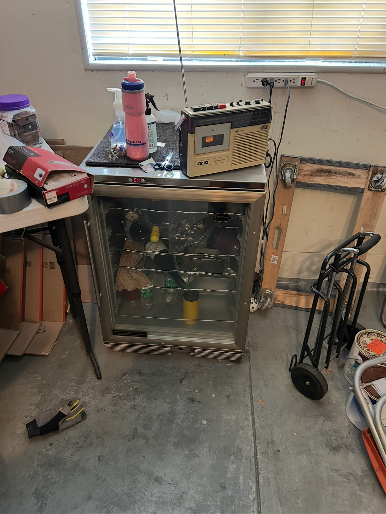 A small refrigerator with a glass door and metal racks inside, a radio on top, surrounded by various items including a water bottle, scissors, and cleaning supplies in a cluttered space.