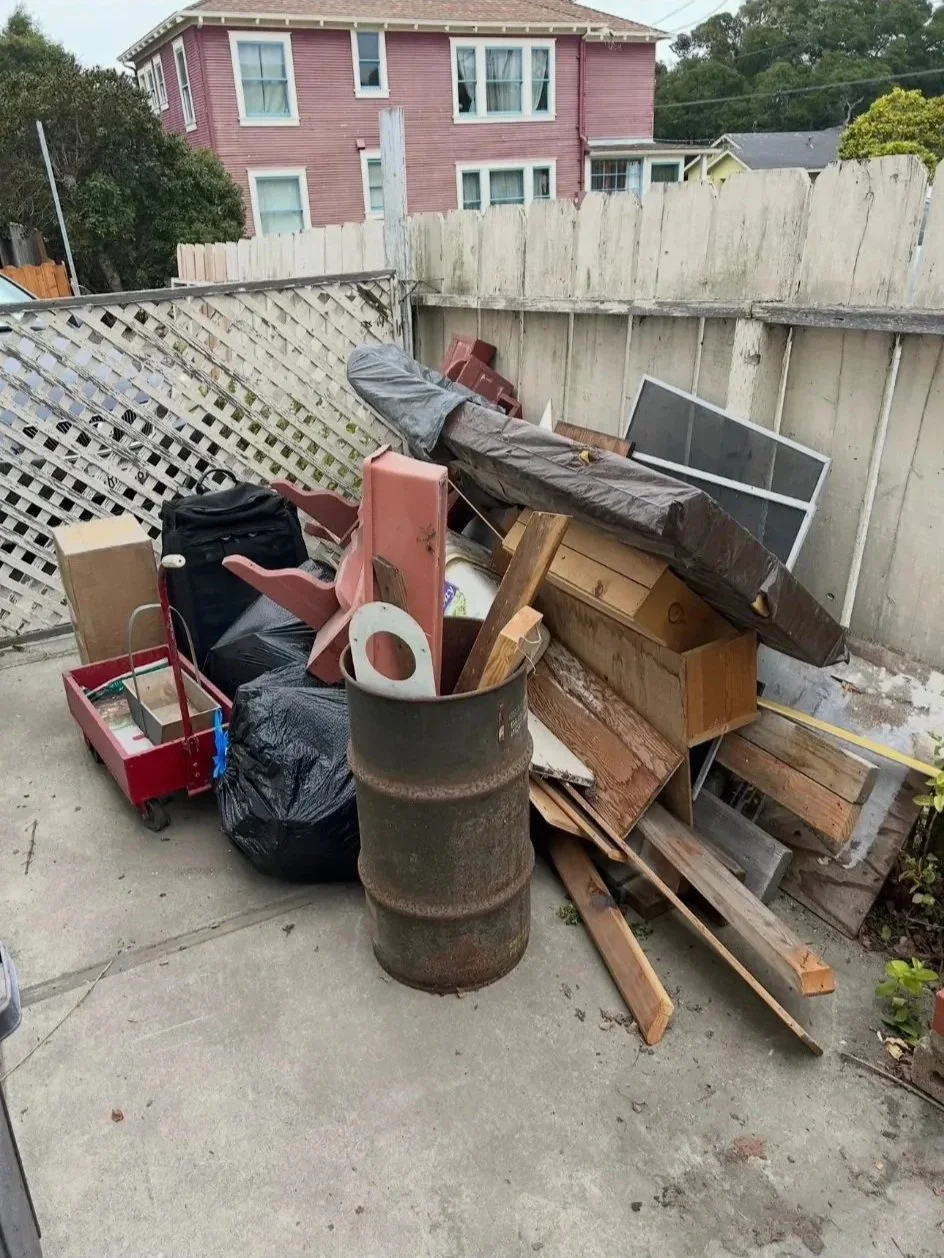 Overflowing backyard with pile of various discarded items including wooden planks, black trash bag, plastic containers, a rusty barrel, a small red wagon, and a piece of furniture, surrounded by a weathered wooden fence, with a pink house in the back