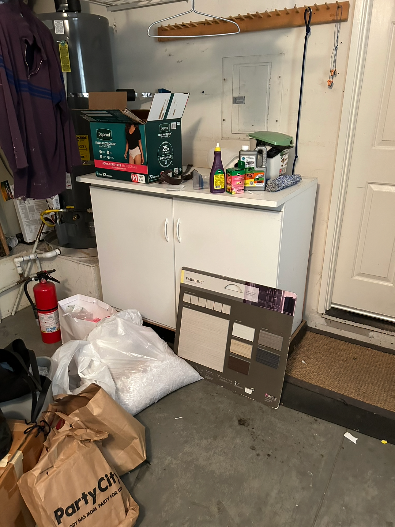Garage corner with a white cabinet covered with cleaning supplies and a box of disposable bed liners, a fire extinguisher on the floor, and various bags and boxes. Items include a water heater, a hose, and a display sample board with paint or floorin