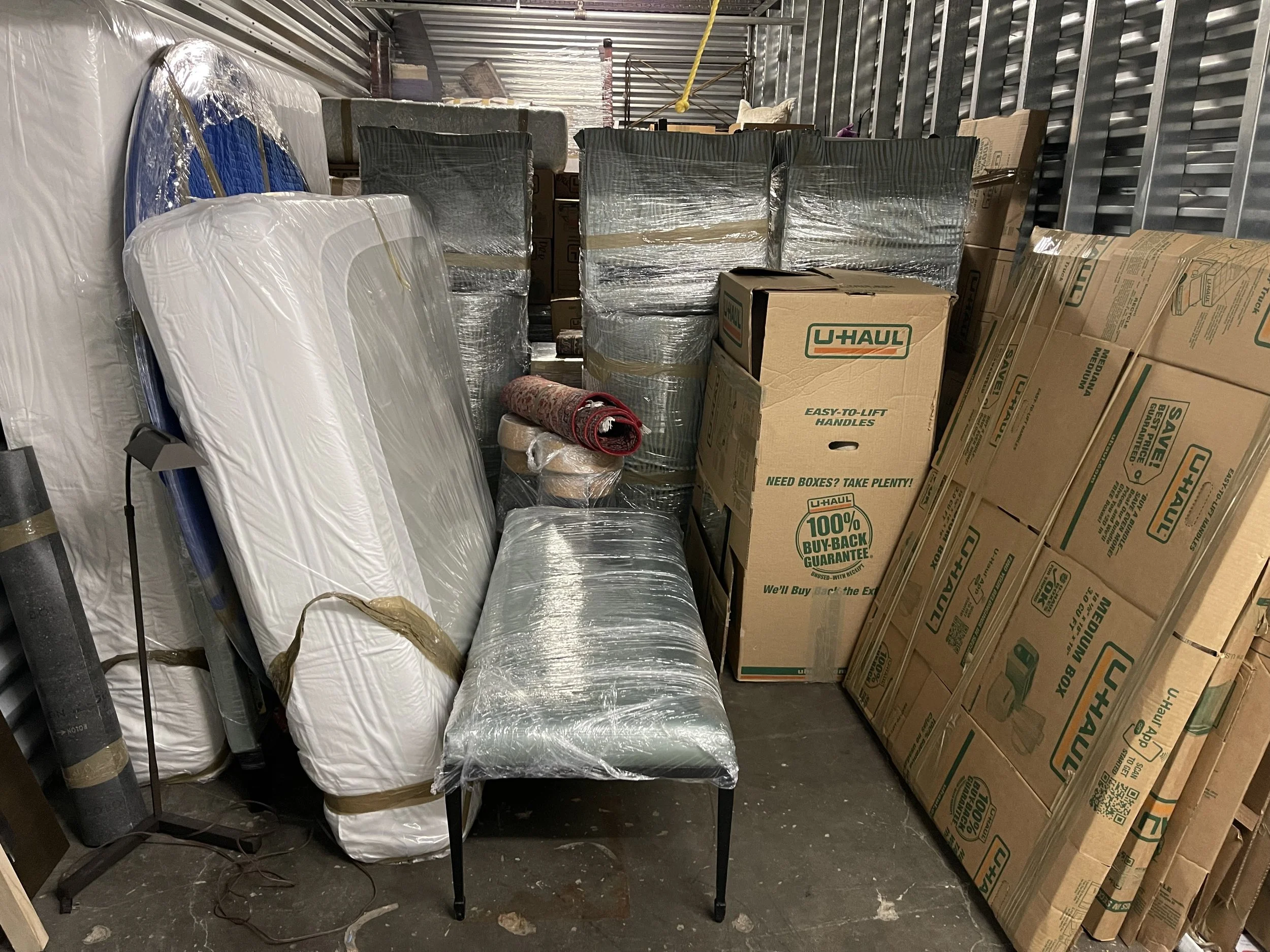 Storage unit filled with boxed and wrapped furniture and boxes, including mattresses, cushions, and moving supplies.