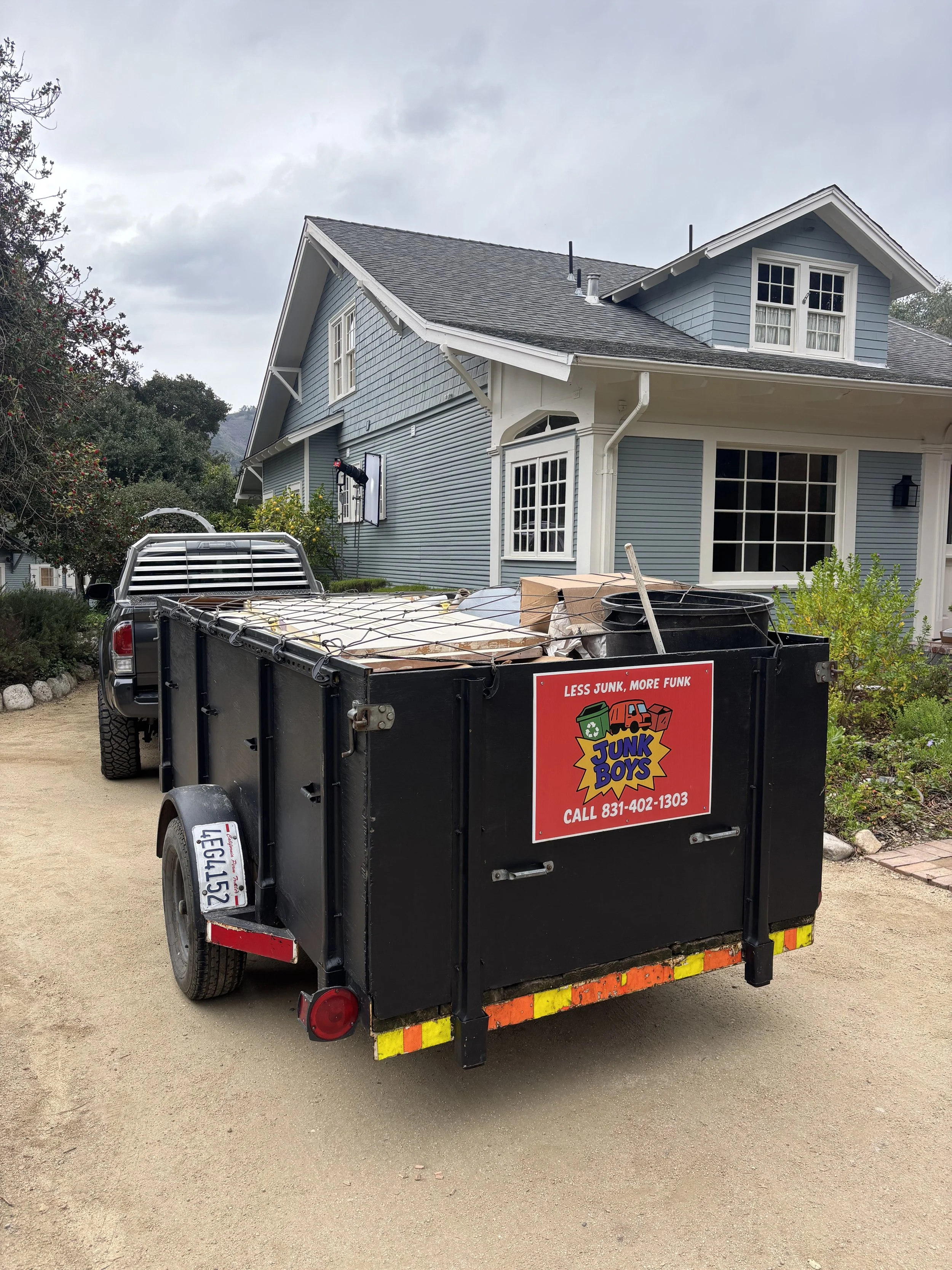 A black trailer attached to a pickup truck, filled with junk, parked on a dirt driveway in front of a blue two-story house with white trim. There is a logo on the trailer that reads 'Junk Boys' with the slogan 'Less Junk, More Funk' and a phone numbe