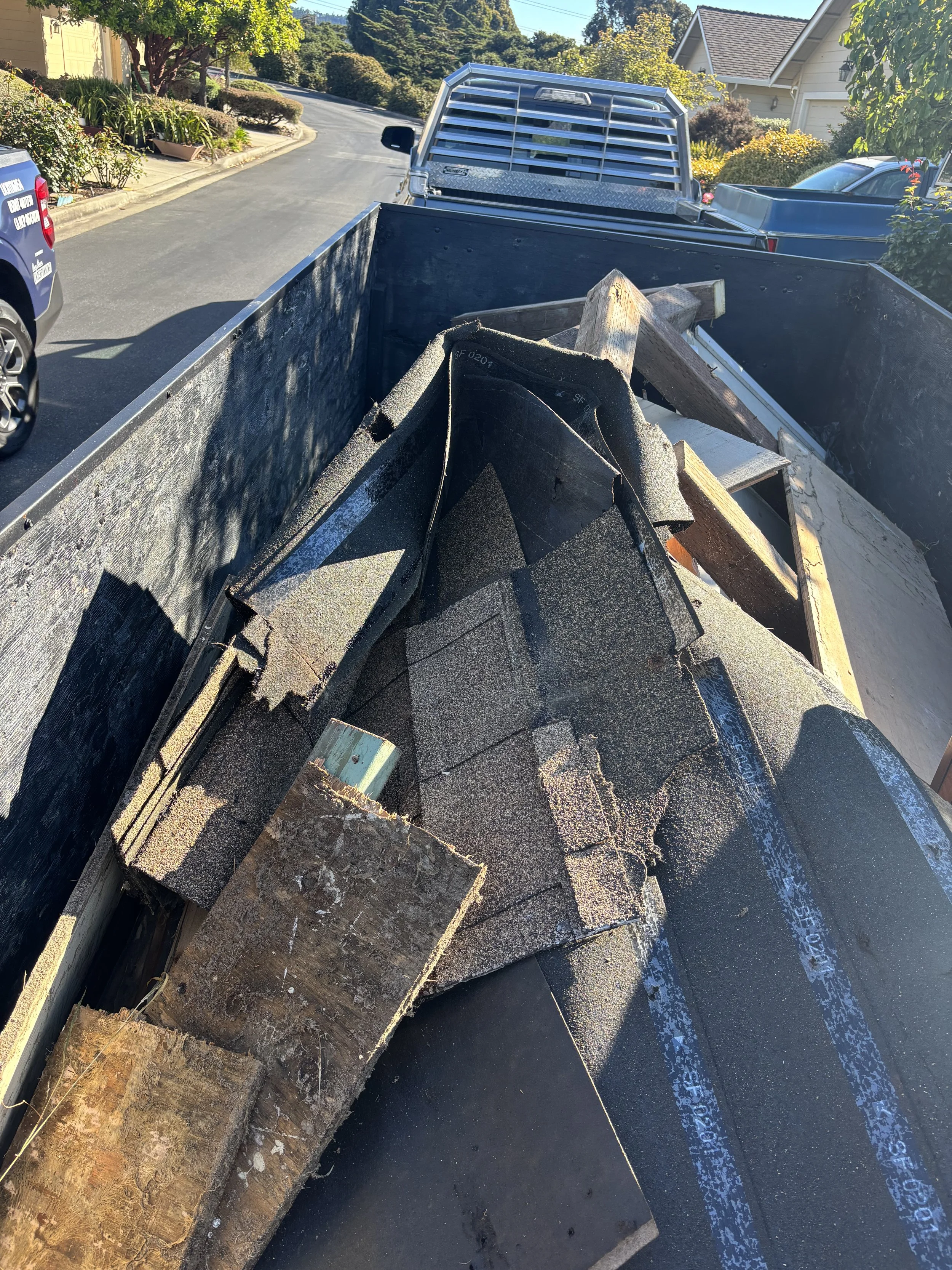 Pickup truck bed filled with scrap construction materials, including wood and roofing shingles, parked on a residential street.