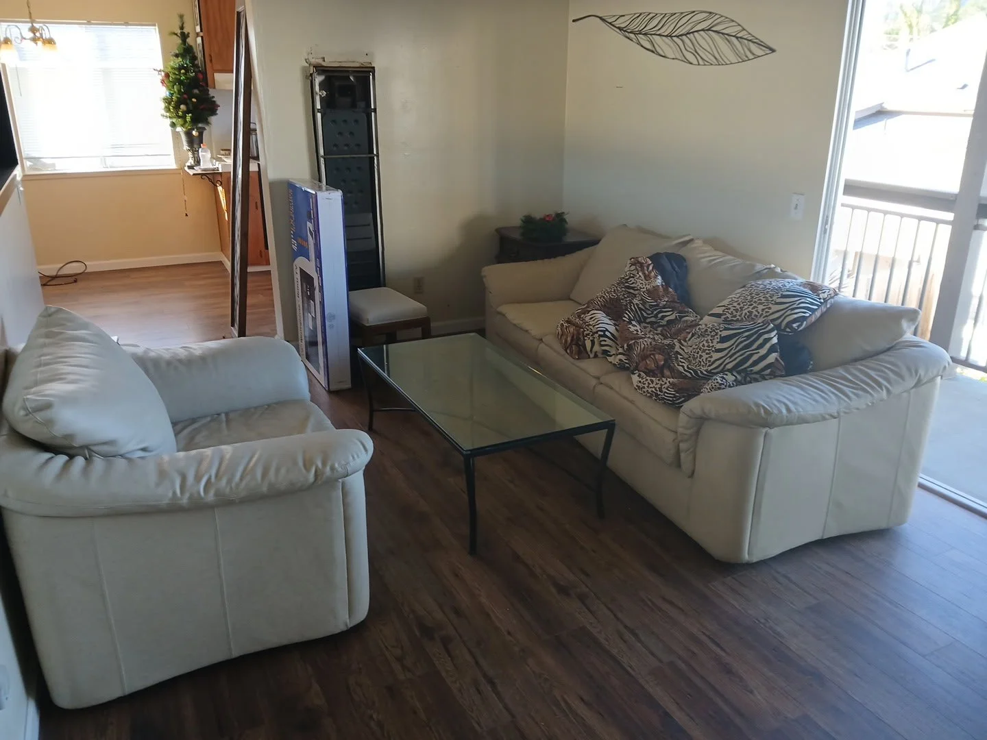 Living room with two white sofas, one with colorful animal print blankets, a glass coffee table, a small side table, a Christmas tree in the corner, and a sliding door leading outside.