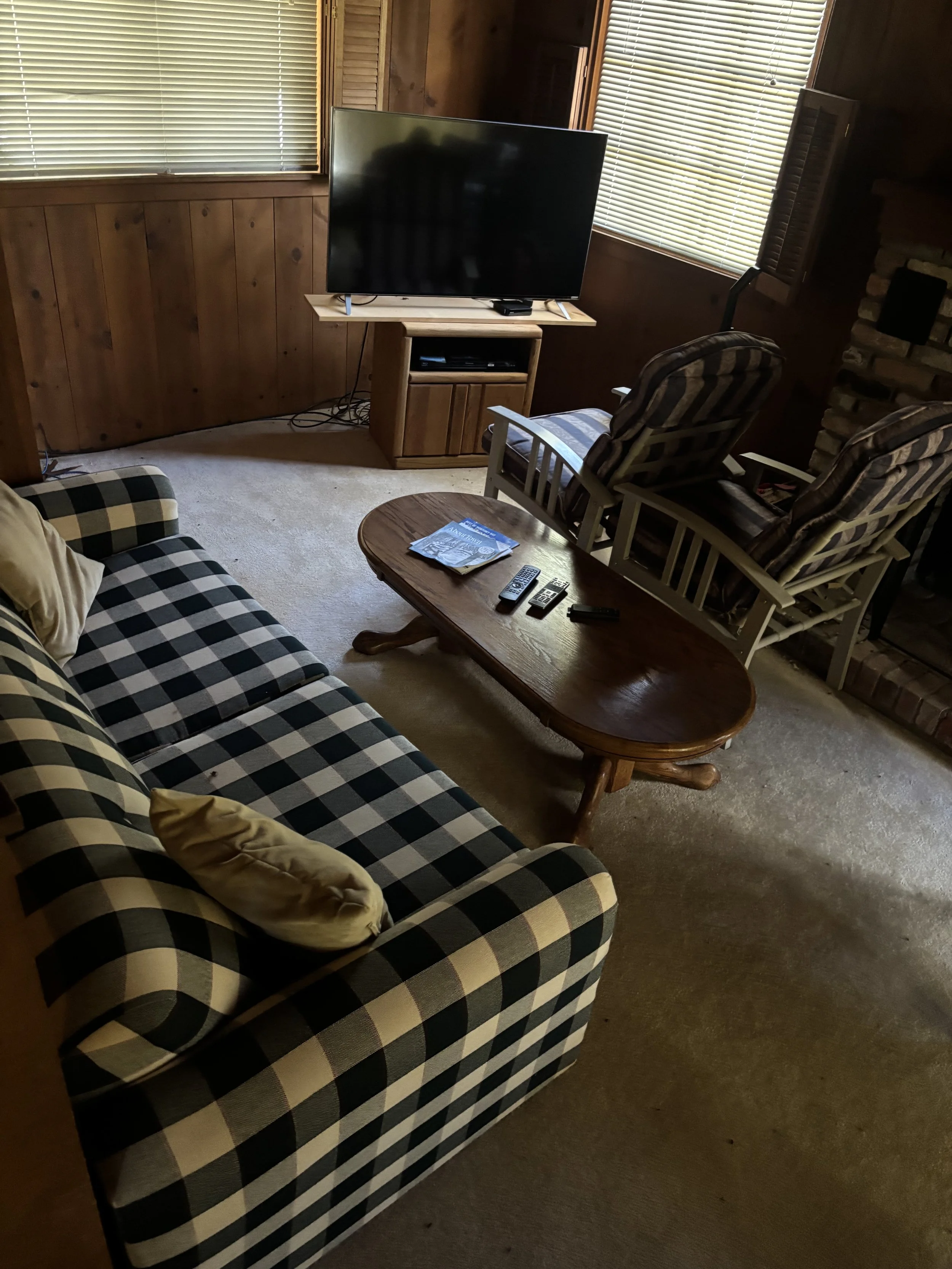 Living room with a checkered sofa, a wooden coffee table with remote controls, a wooden TV stand with a large flat screen TV, two armchairs with striped cushions, and large windows with blinds.