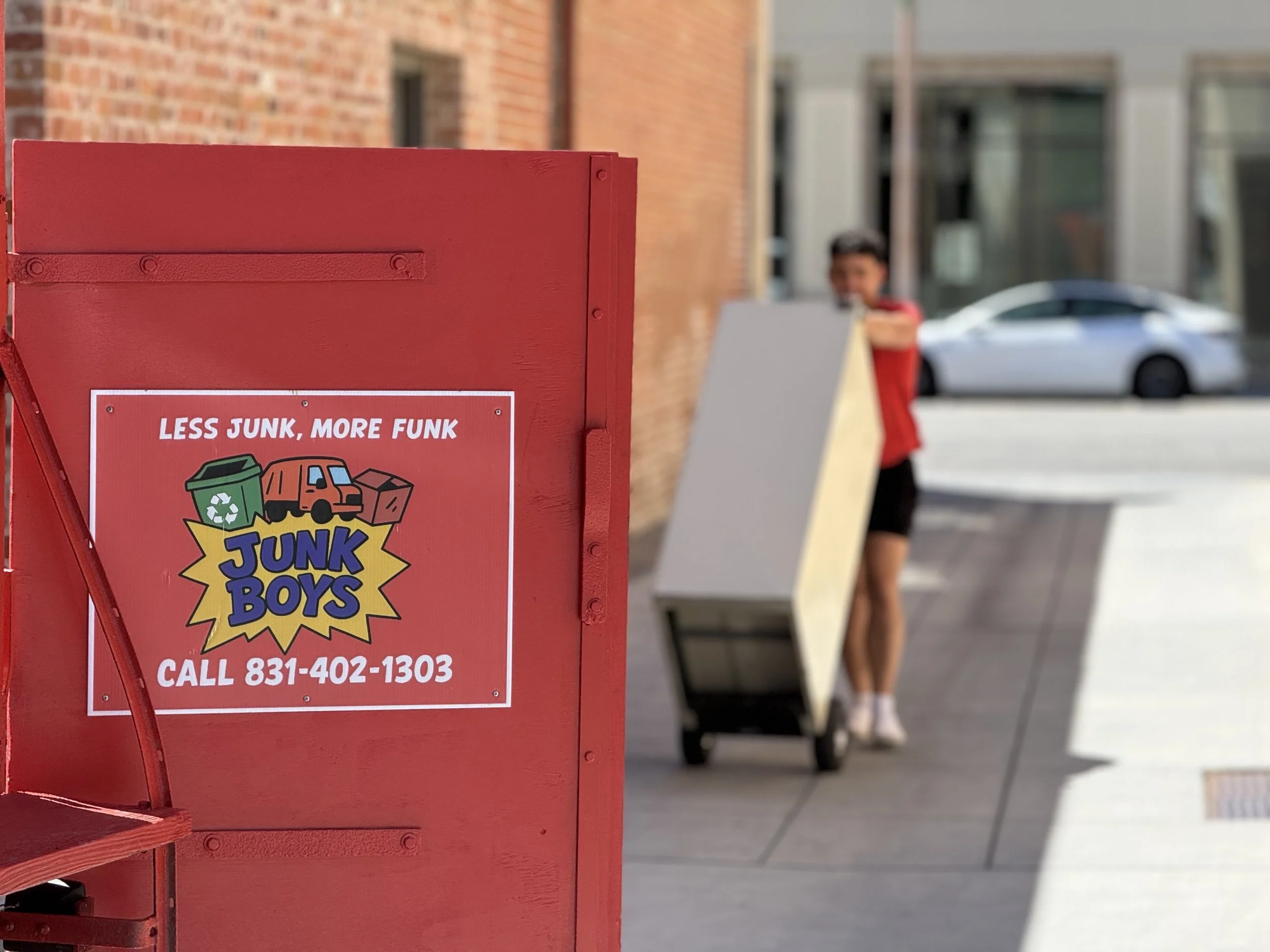 Close-up of a red recycling bin with a label that reads "Less Junk, More Funk" and a phone number. In the blurred background, a person in a red shirt is walking along the sidewalk, with parked cars and buildings visible.