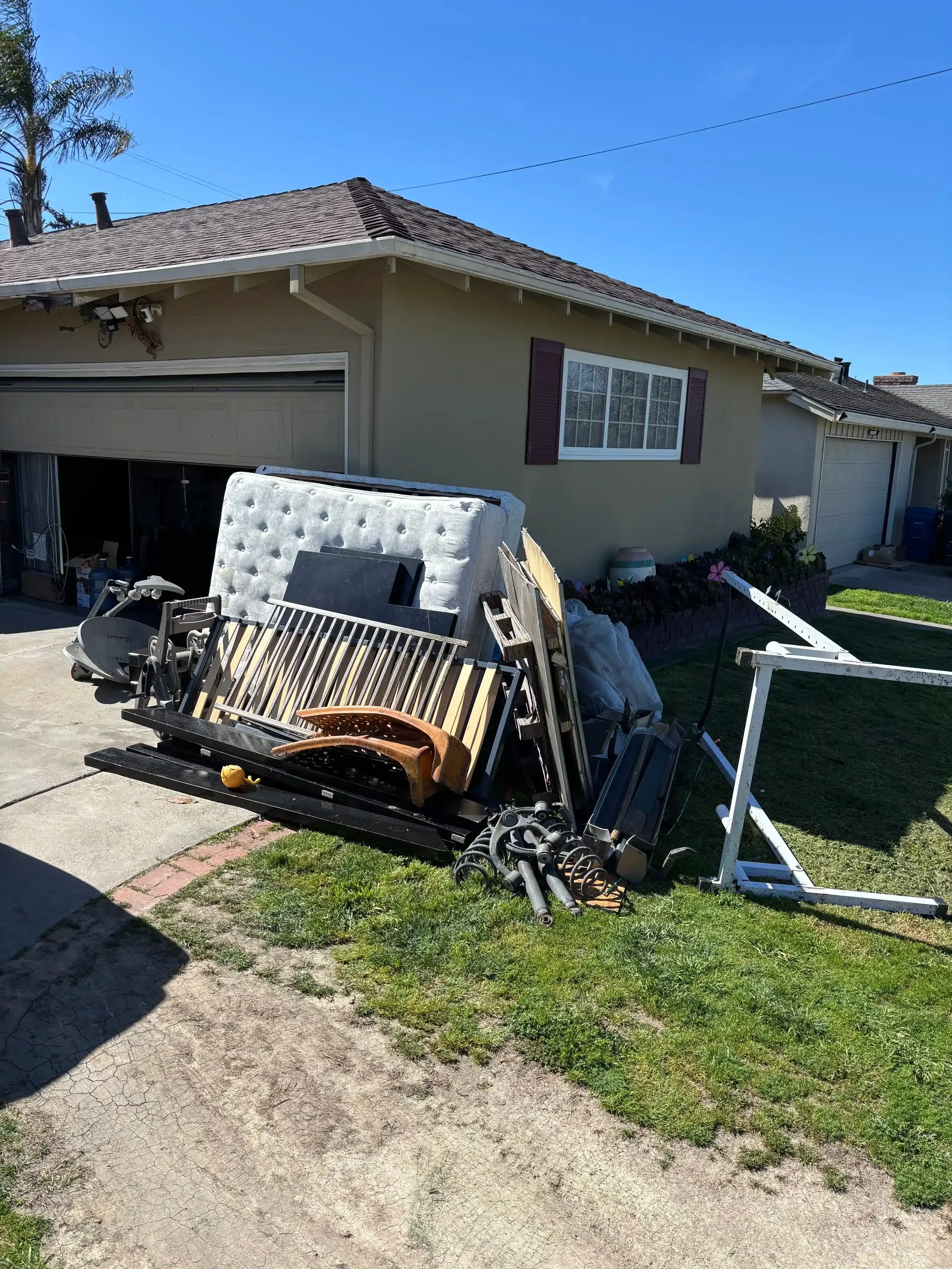 A pile of discarded furniture and household items in front of a house, including a mattress, bed frame, office chair, and various metal parts. Salinas Junk removal.