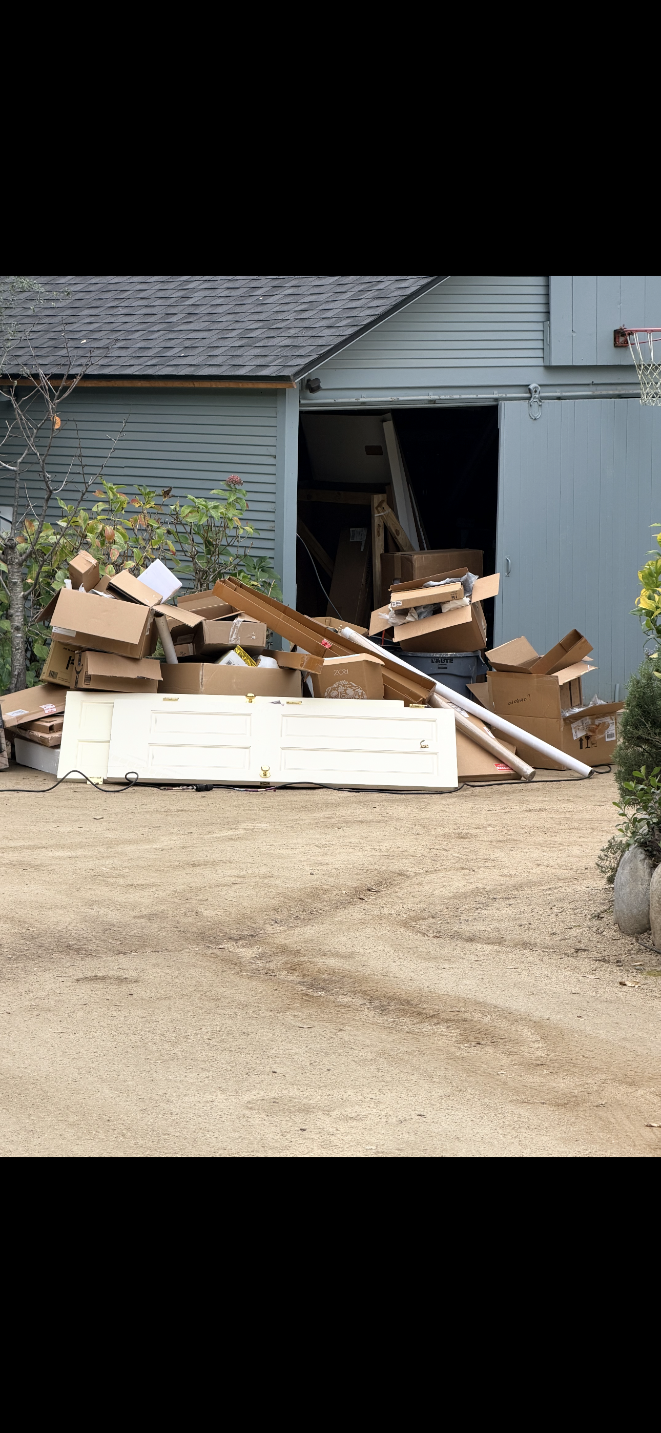 Piles of cardboard boxes, a white door, and miscellaneous items outside a blue garage or shed with a basketball hoop attached.