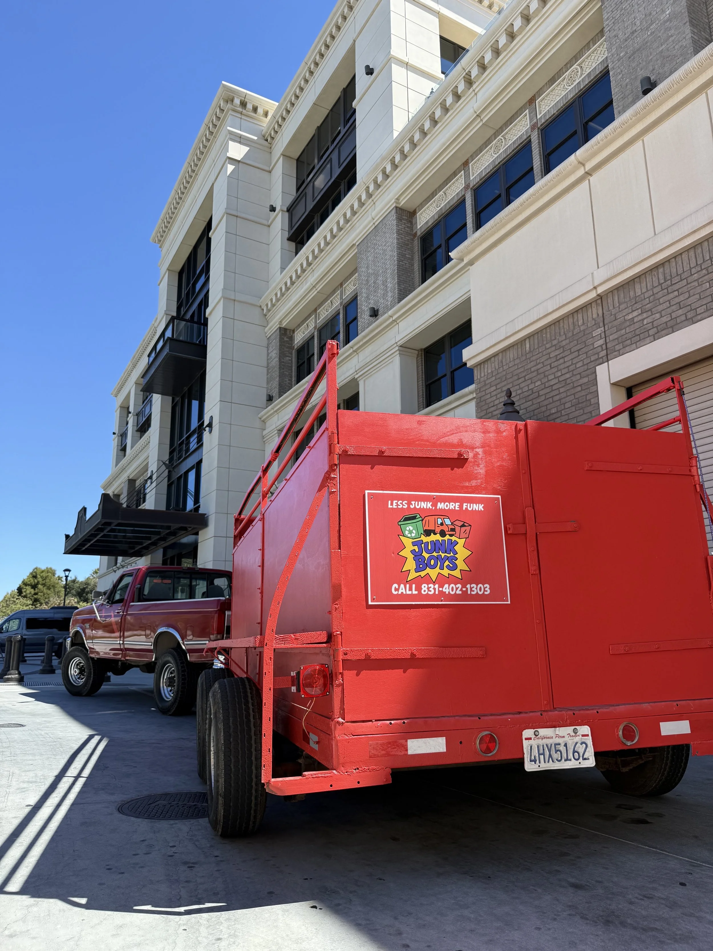 Red junk removal truck with a logo reading 'Junk Boys' parked on a city street in front of modern buildings with clear blue sky overhead.