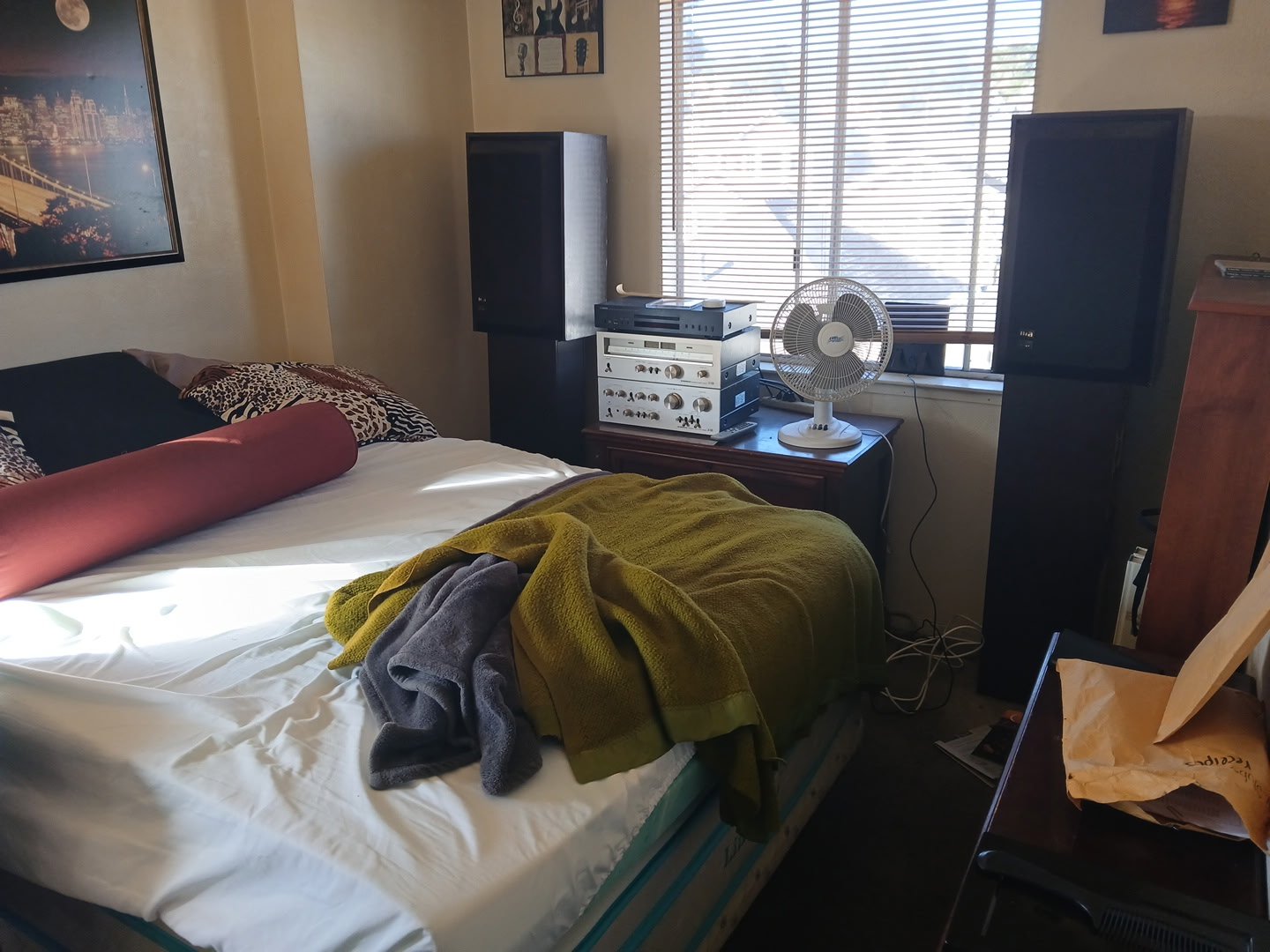 A bedroom with a bed, a window with blinds, a stereo system with a fan on top, and some furniture. Clothes and a bolster are on the bed, and a paper bag is on a table.