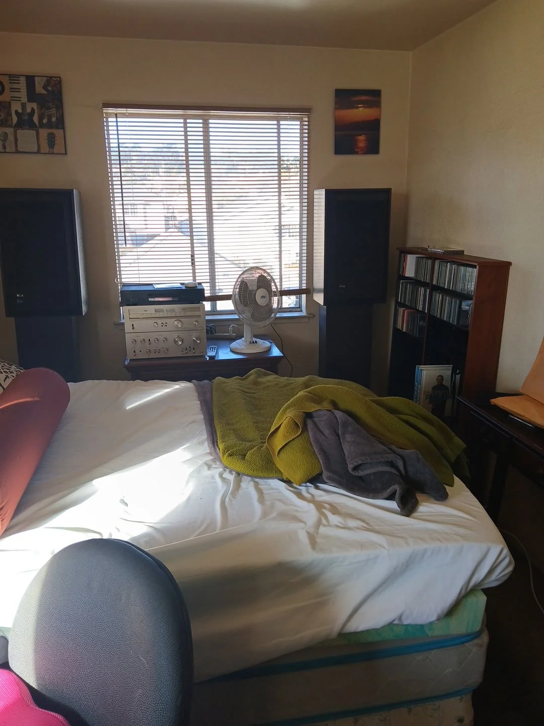 A bedroom with a bed and a window with blinds. The bed has pillows and blankets. There is a fan and stereo system on a table by the window. A picture hangs on the wall, and a bookcase with CDs is near the bed.
