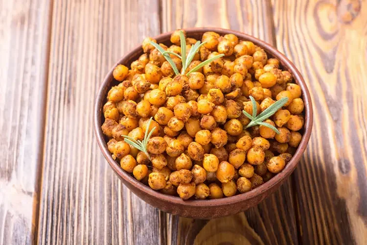 A wooden bowl filled with golden-brown roasted chickpeas seasoned with rosemary. This crunchy, high-protein snack is an excellent plant-based alternative to traditional chips, helping support digestion.