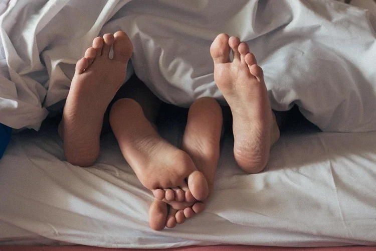 A low-section view of a couple's feet peeking out from under a white blanket, symbolizing physical closeness and deep relaxation.