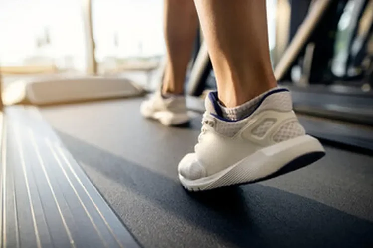 A close-up shot of a man's feet running on a treadmill belt in an indoor gym. It highlights the controlled environment and the shock-absorbing surface of treadmill running.