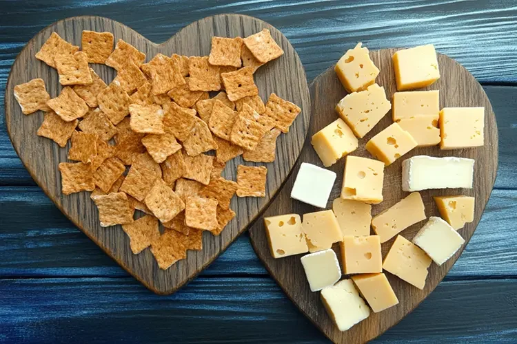 Two heart-shaped wooden boards featuring whole-grain crackers and cubed cheese pieces, set against a dark blue background. It represents a balanced snack that combines protein and complex carbs to keep you focused.