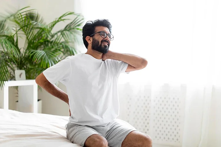A man sitting on a bed holding his neck and lower back in discomfort, illustrating common morning stiffness in winter.