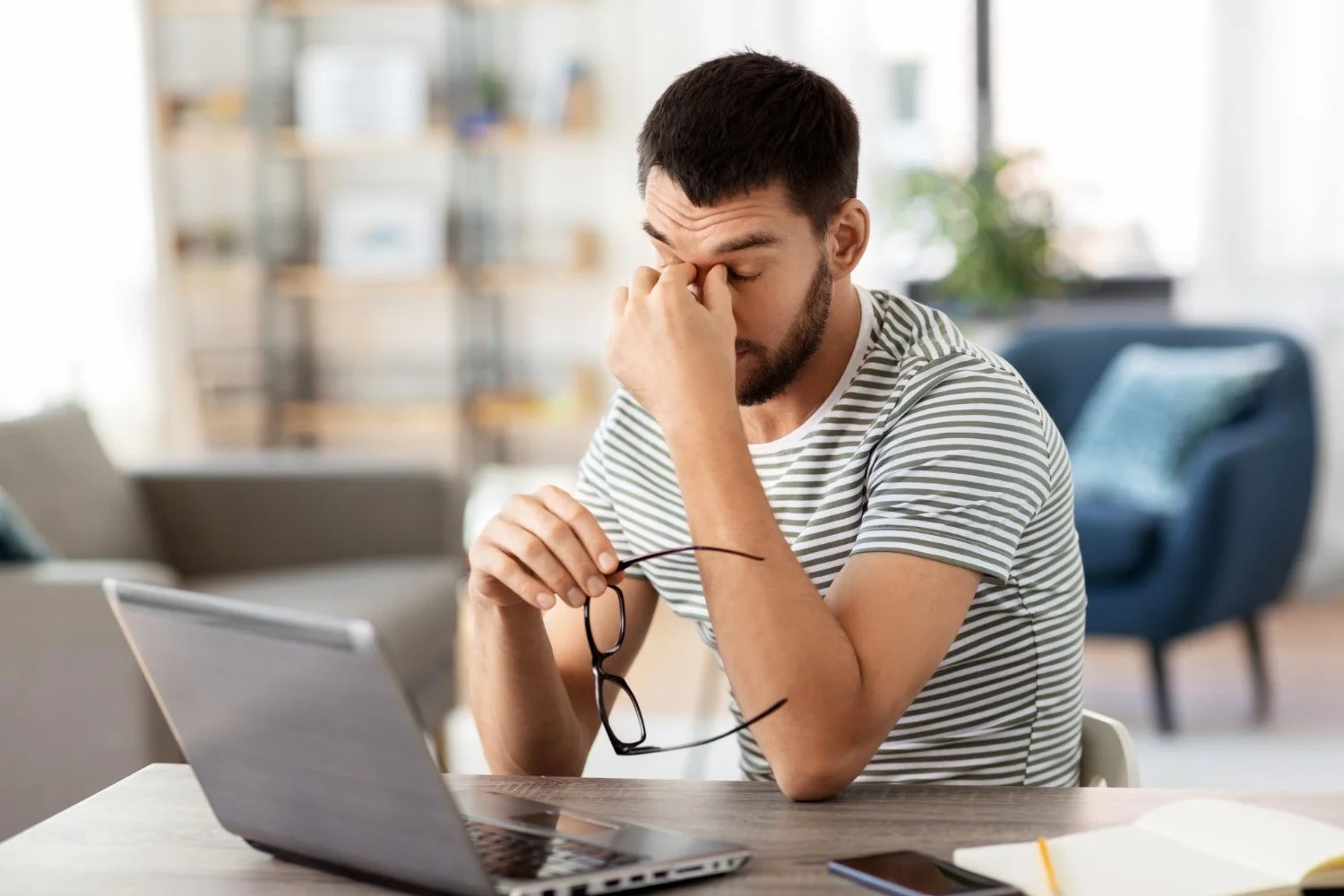 Tired man rubbing his eyes while working on a laptop at a home office desk, showing signs of stress and fatigue.