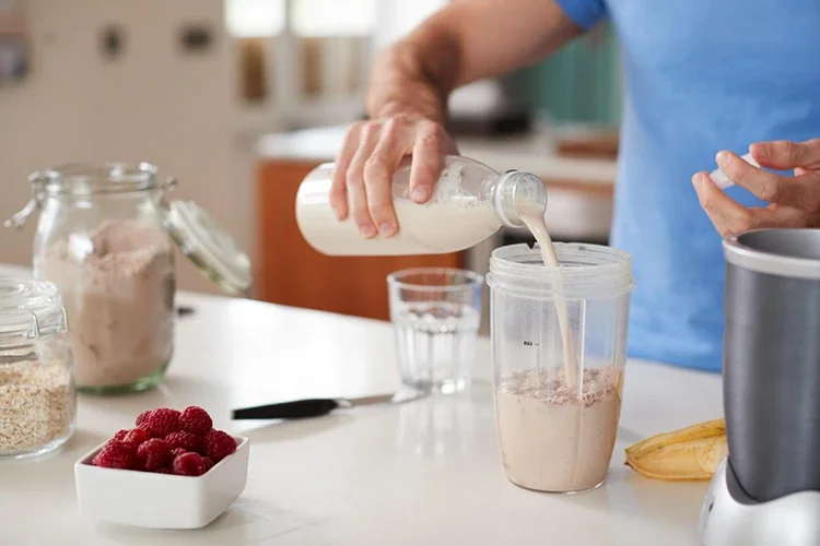 A close-up of a man pouring liquid into a blender cup in a kitchen. Ingredients like protein powder, raspberries, and a banana are visible on the counter, illustrating the process of preparing a supplement-based drink.