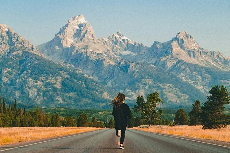 A woman running along an open road with scenic mountains in the background. It emphasizes the importance of outdoor training for adapting to real-world terrain and race conditions.