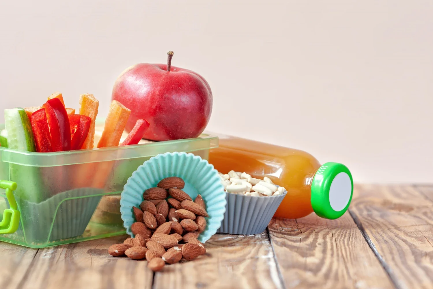 A vibrant shot featuring a transparent lunch box filled with fresh veggie sticks (carrots and peppers), a whole red apple, a cup of almonds, and a bottle of orange juice on a wooden surface.