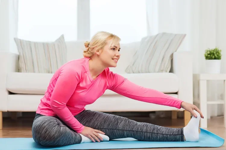 A happy woman smiling while stretching her leg on a yoga mat at home, promoting body positivity and a healthy lifestyle.