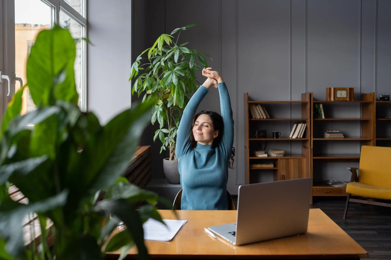 A woman takes a mindful break from working on her laptop, stretching and gazing out the window in a shared office space