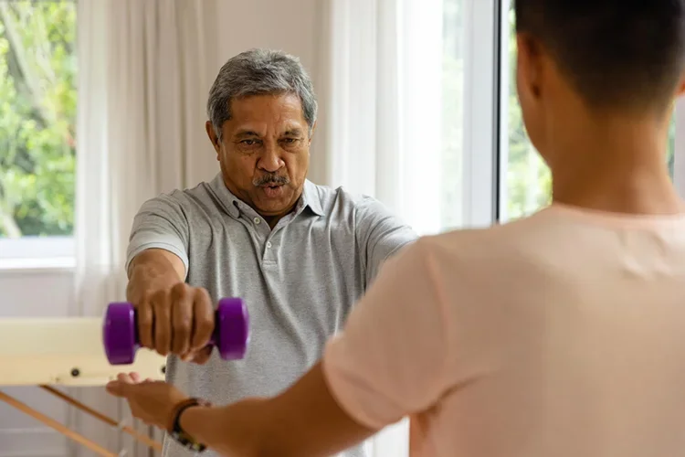 A senior male patient performing rehabilitation exercises with dumbbells under the guidance of a male physiotherapist. It highlights the role of professional physical therapy in managing chronic pain.
