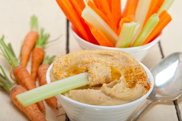 Crunchy carrot and celery sticks paired with a creamy hummus dip in a white bowl. This combination represents a satisfying, plant-based snack rich in fiber and essential vitamins, perfect for weight management.