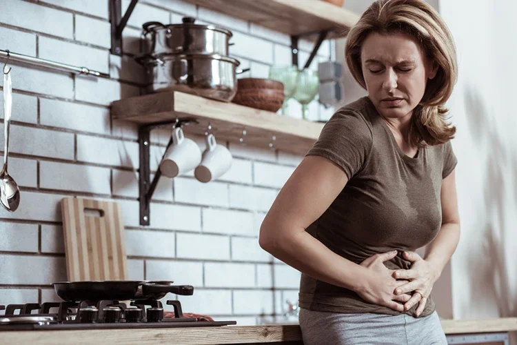 A blonde woman in a kitchen holding her stomach with a pained expression. It symbolizes potential digestive distress or side effects resulting from the nutritional gaps of relying solely on supplements as meal replacements.