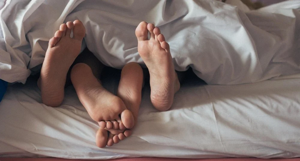 Close-up of a couple’s legs intertwined under a blanket in bed, symbolizing physical intimacy, comfort, and the healing power of closeness.
