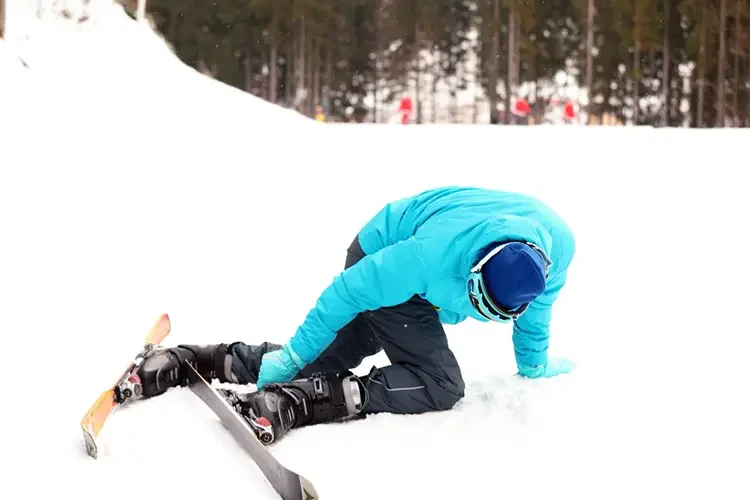 A male skier in a blue jacket fallen on the snow with skis tangled, illustrating how twisting forces lead to knee ligament injuries.