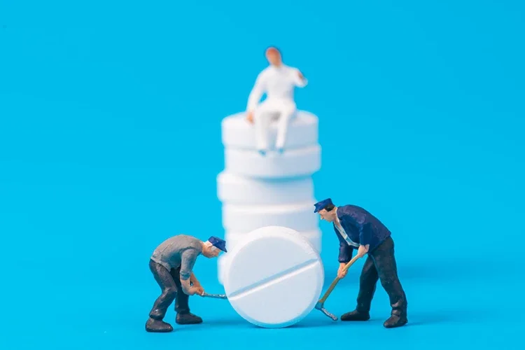 Close-up of figurines surrounded by various medicines on a blue background, symbolizing increasing dependence on medication to mask pain.