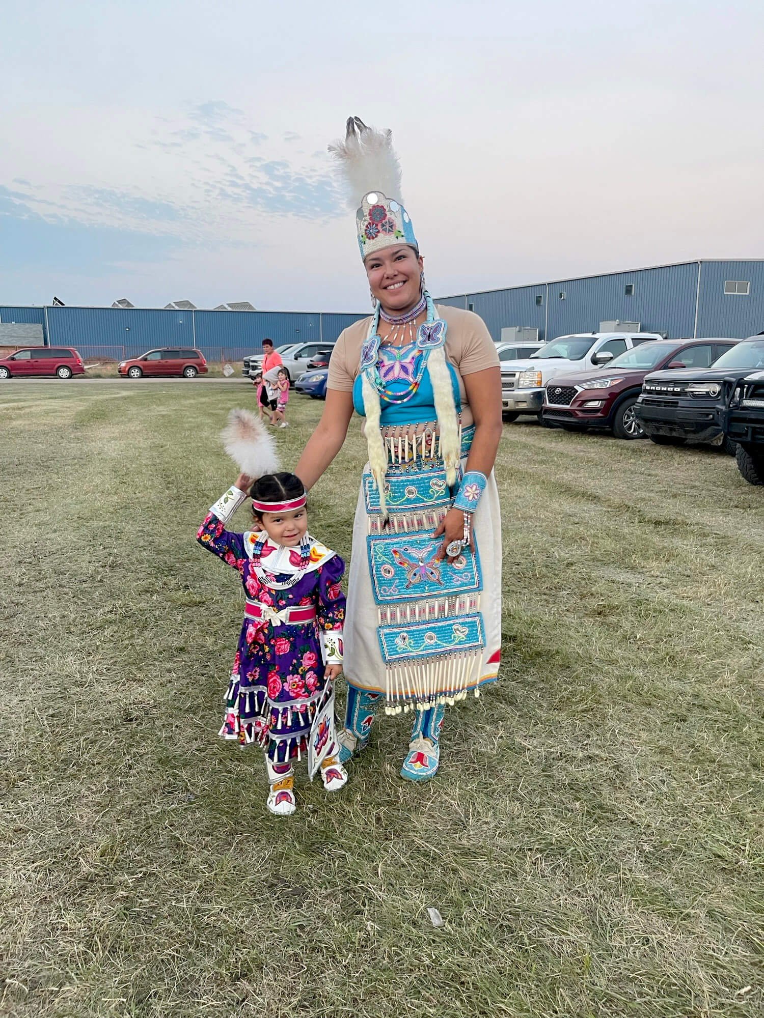 Two women dressed in traditional Native American regalia, standing outdoors on a grassy field with cars and a blue building in the background. The woman and girl are smiling, wearing colorful beaded clothing, feathered headdresses, and accessories.